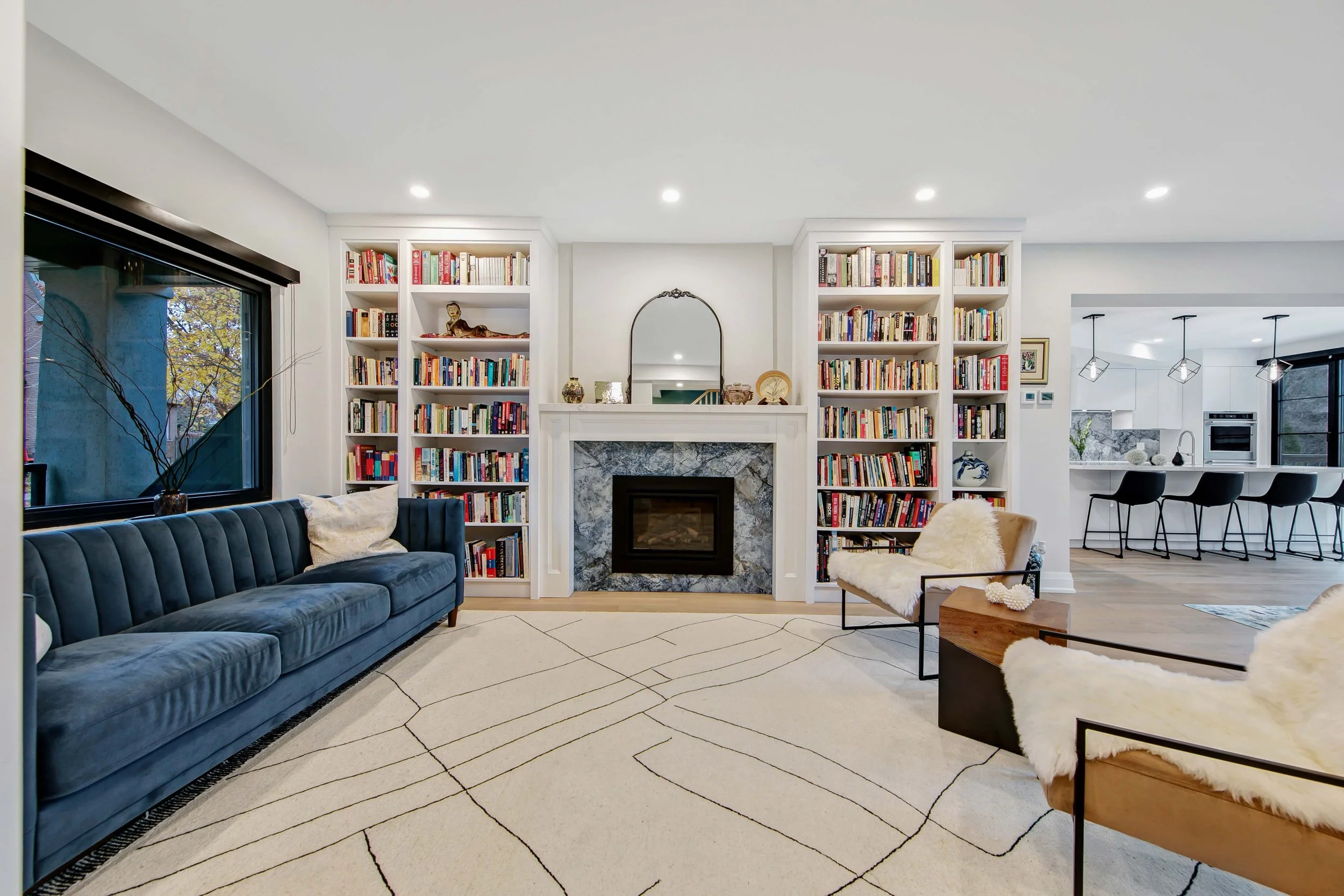 Living room with a dark blue velvet sofa, white patterned rug, built-in white bookshelves with books and decorative items, a marble fireplace, and a large window revealing trees outside. Kitchen with white cabinets, black pendant lights, and barstool