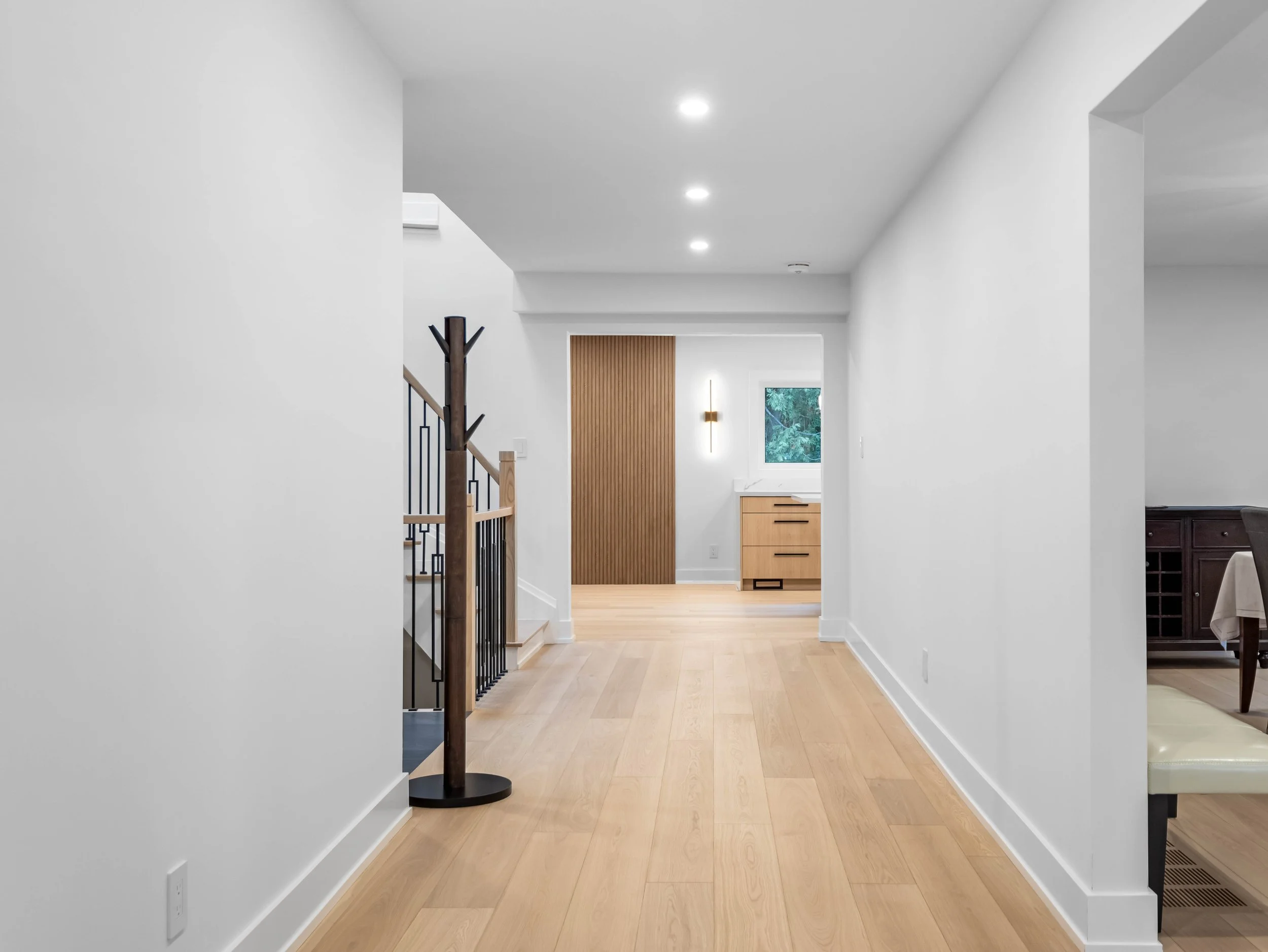 A modern, minimalist home interior with white walls, wooden flooring, a staircase with black metal railing and wooden handrail, and a view toward a hallway with natural light through a window. There are some built-in wooden cabinets and a wall-mounte