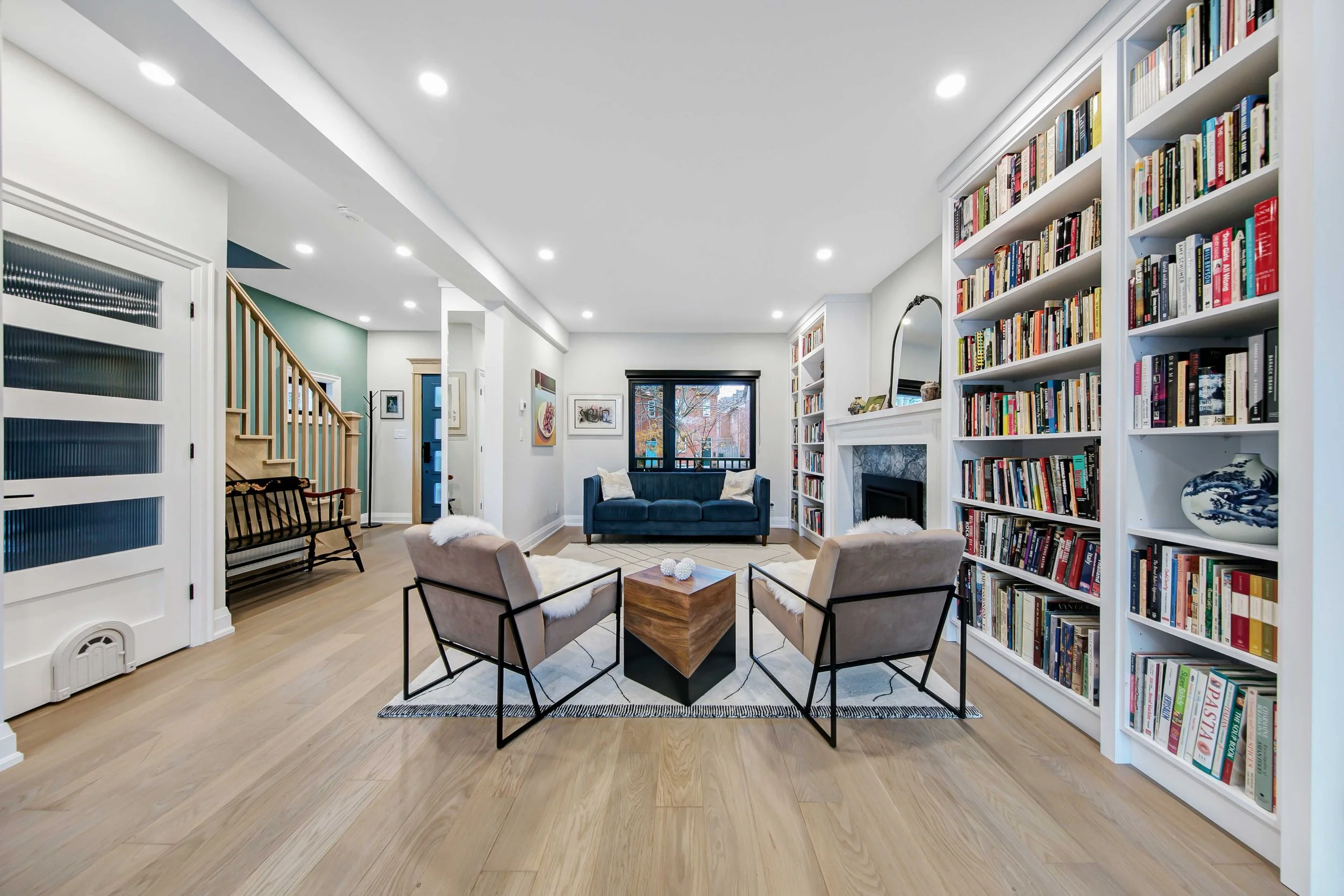 Living room with white walls, wood flooring, a navy sofa, beige chairs around a wooden coffee table, bookshelves filled with books, and artwork on the walls.