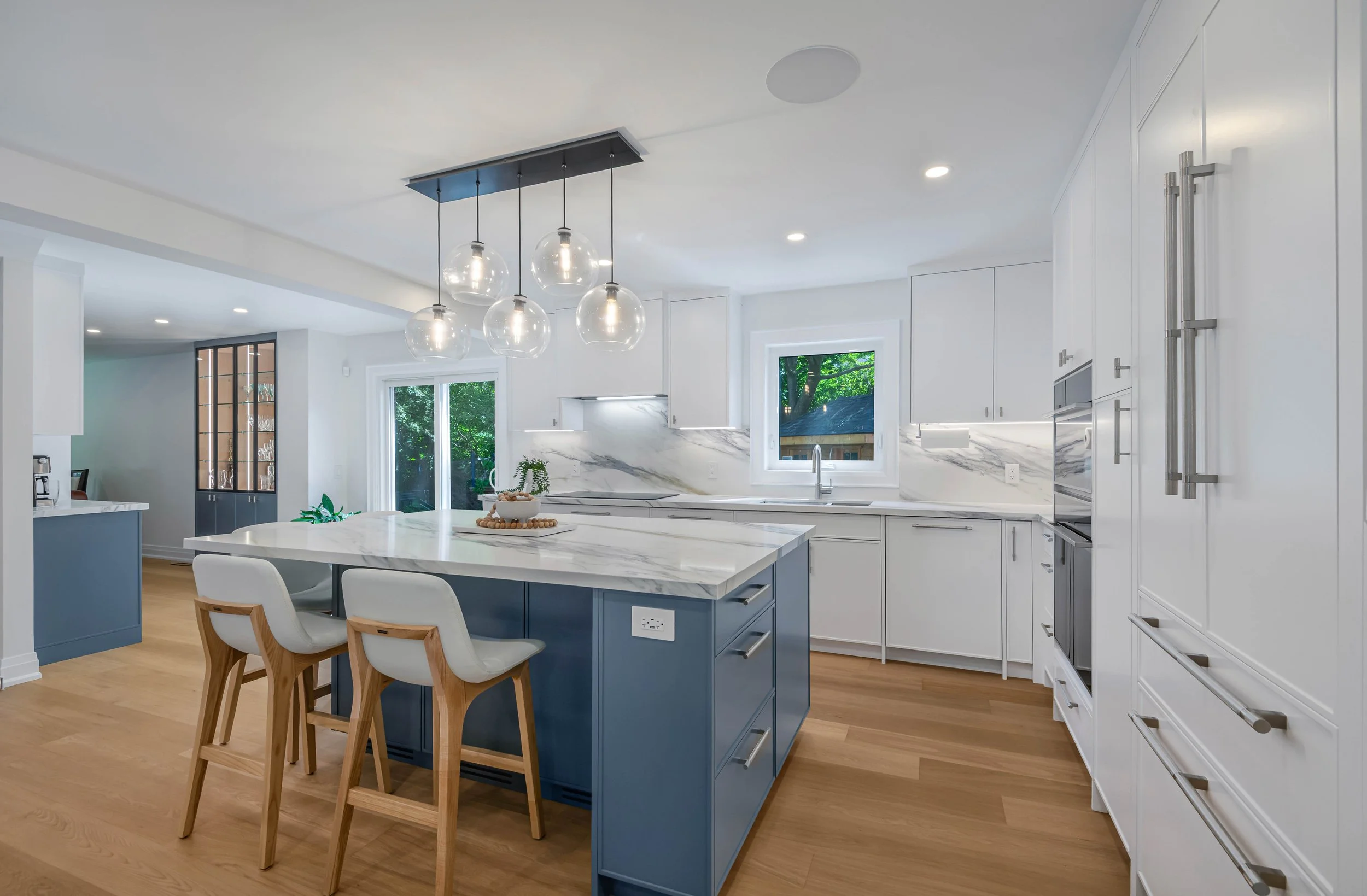 Modern kitchen with white cabinets, marble countertops, and a blue island with wooden bar stools. There is a pendant light fixture with five glass globes hanging above the island. Large windows show green trees outside.