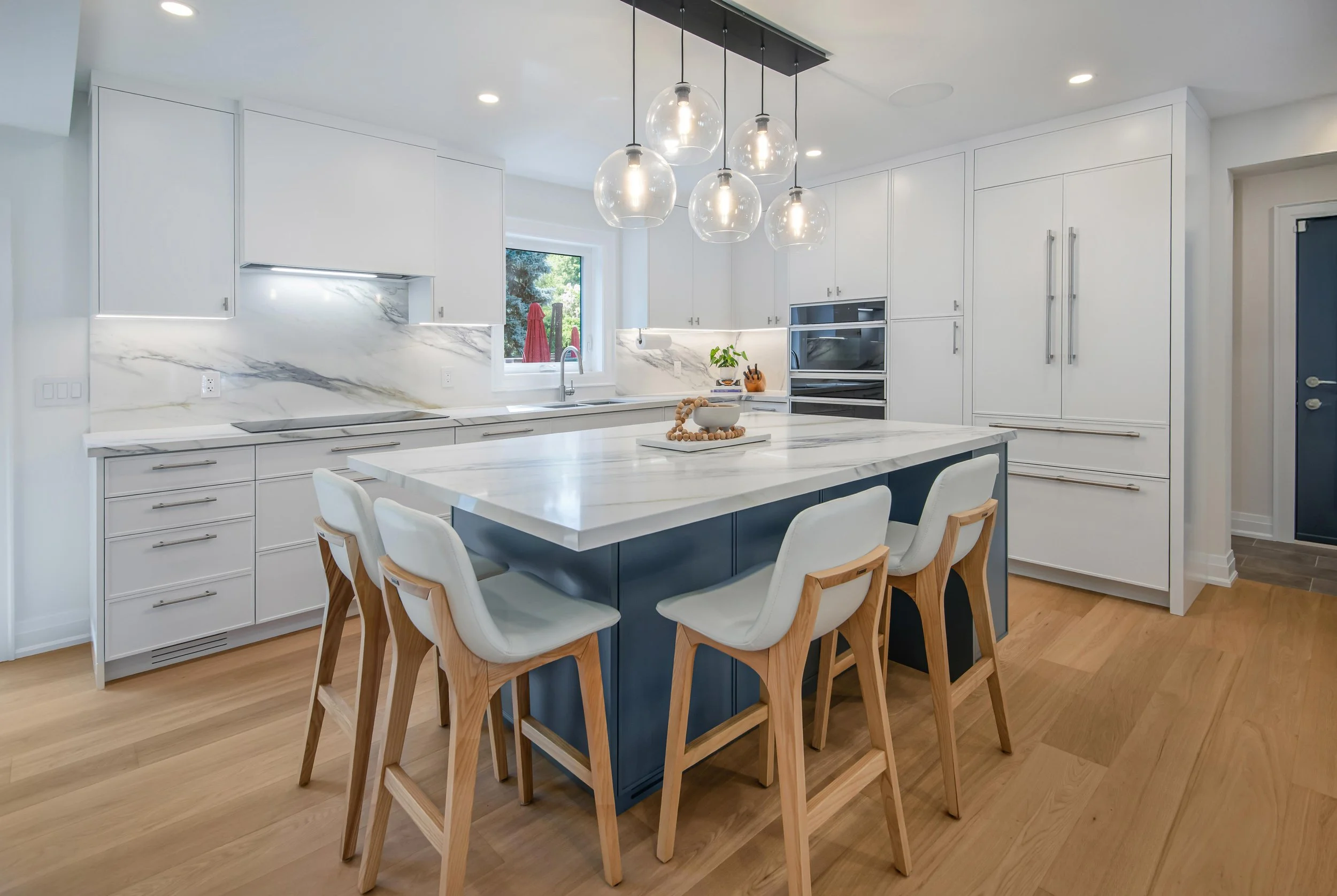 Modern kitchen with white cabinetry, marble countertops, and a central island with blue base. Wooden chairs surround the island. Pendant lights hang above, and a window above the sink shows green outdoor scenery.