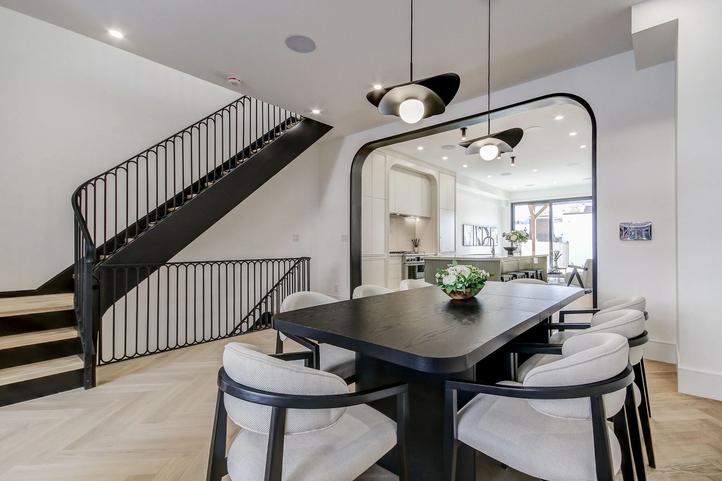 Modern open-concept living space with a dining area, kitchen, and staircase, featuring minimalist black and white decor, pendant lighting, and large windows letting in natural light.