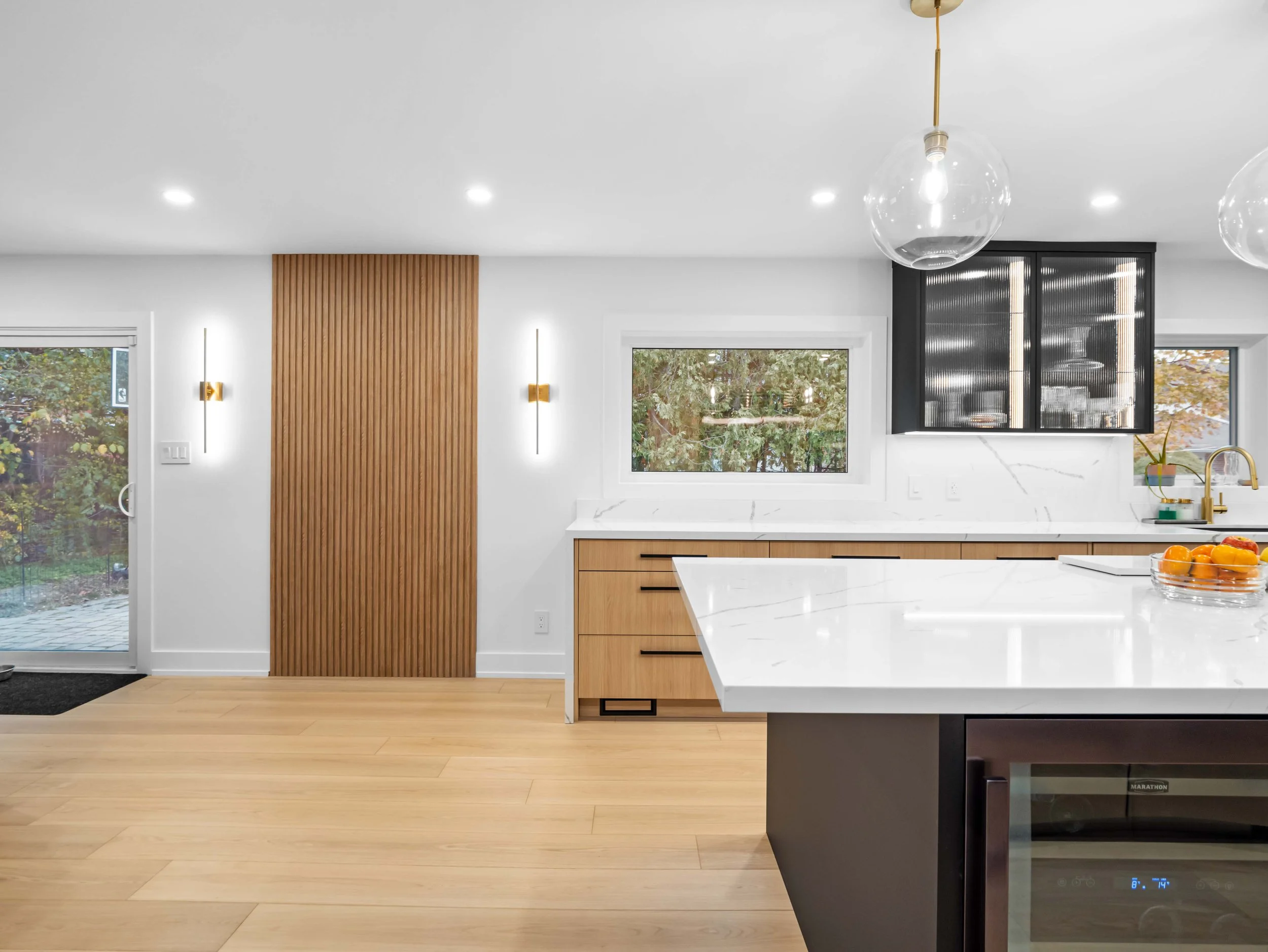 Modern kitchen with white marble island, wooden cabinets, large windows showing outdoor greenery, and contemporary lighting fixtures.