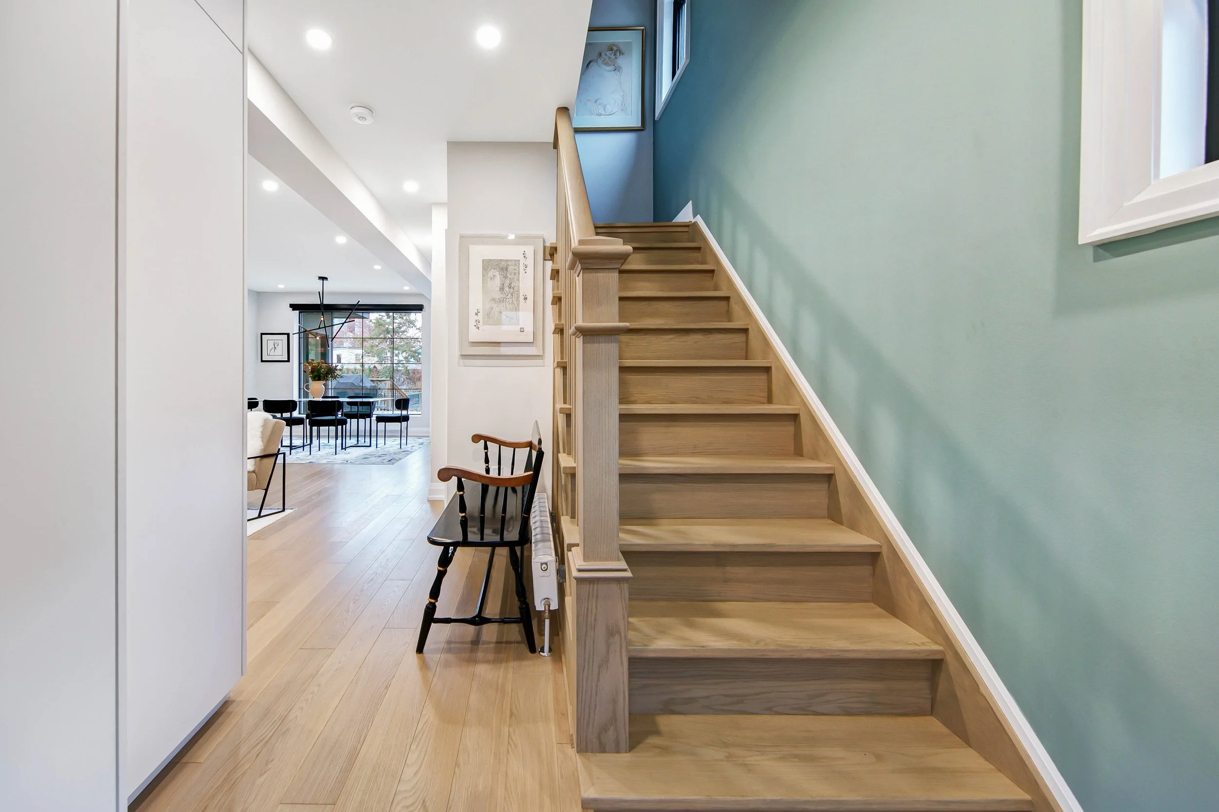 Interior view of a modern home with wooden staircase, light green wall, black chairs, and a kitchen dining area with large window and art on the walls.