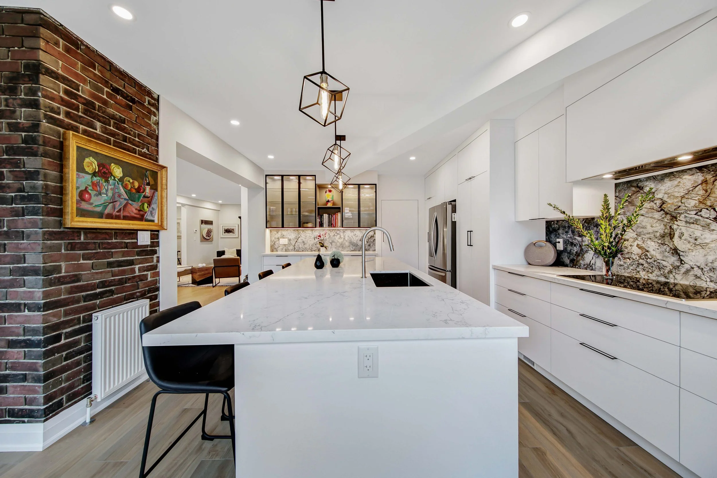 Modern kitchen with white cabinetry, marble countertops, brick accent wall, and pendant lighting. Includes kitchen island with black chairs and a built-in open shelf with books and decorative items. Artwork hangs on brick wall, and there are stainles