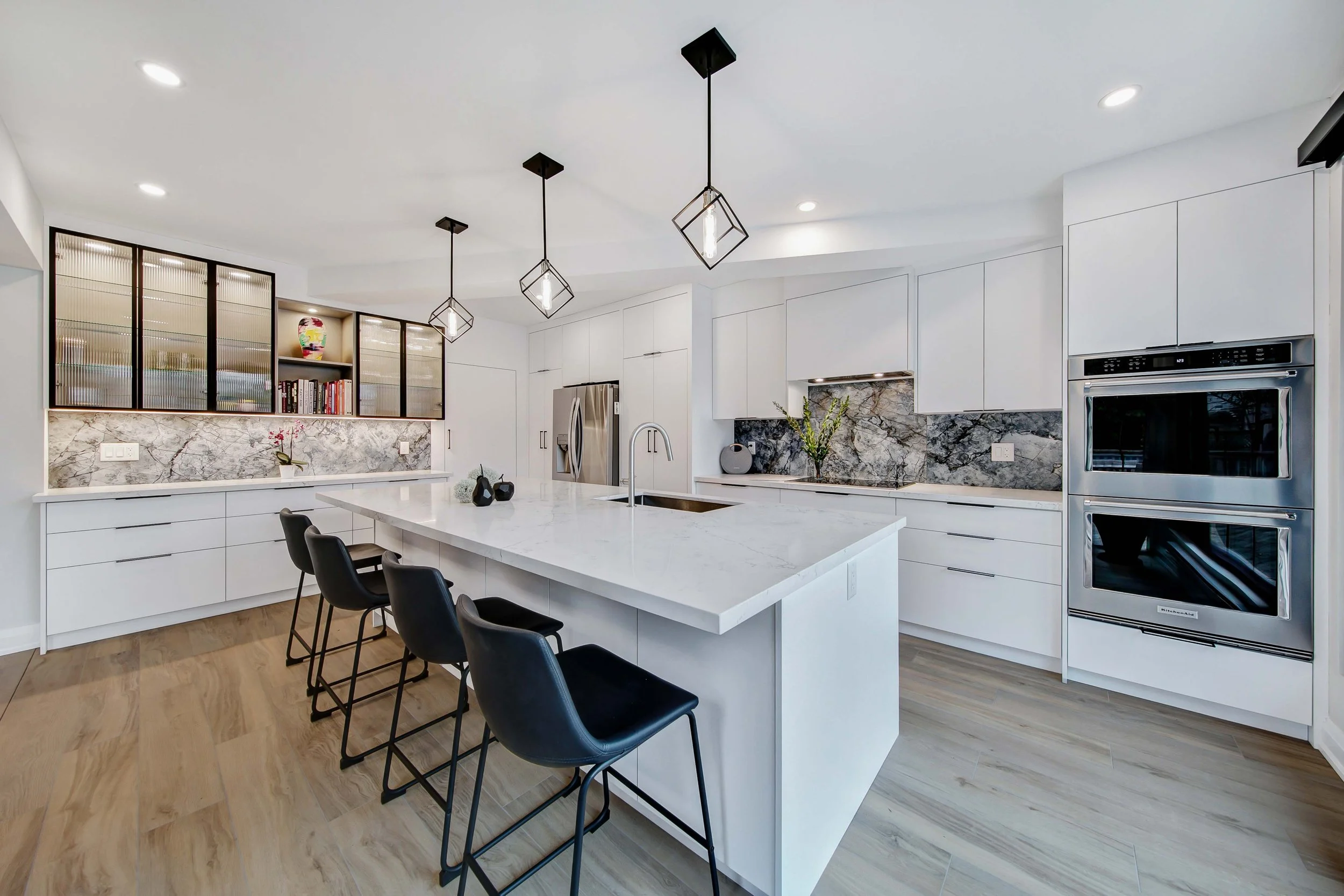 Modern white kitchen with marble backsplash, black pendant lights, black chairs, and stainless steel appliances.