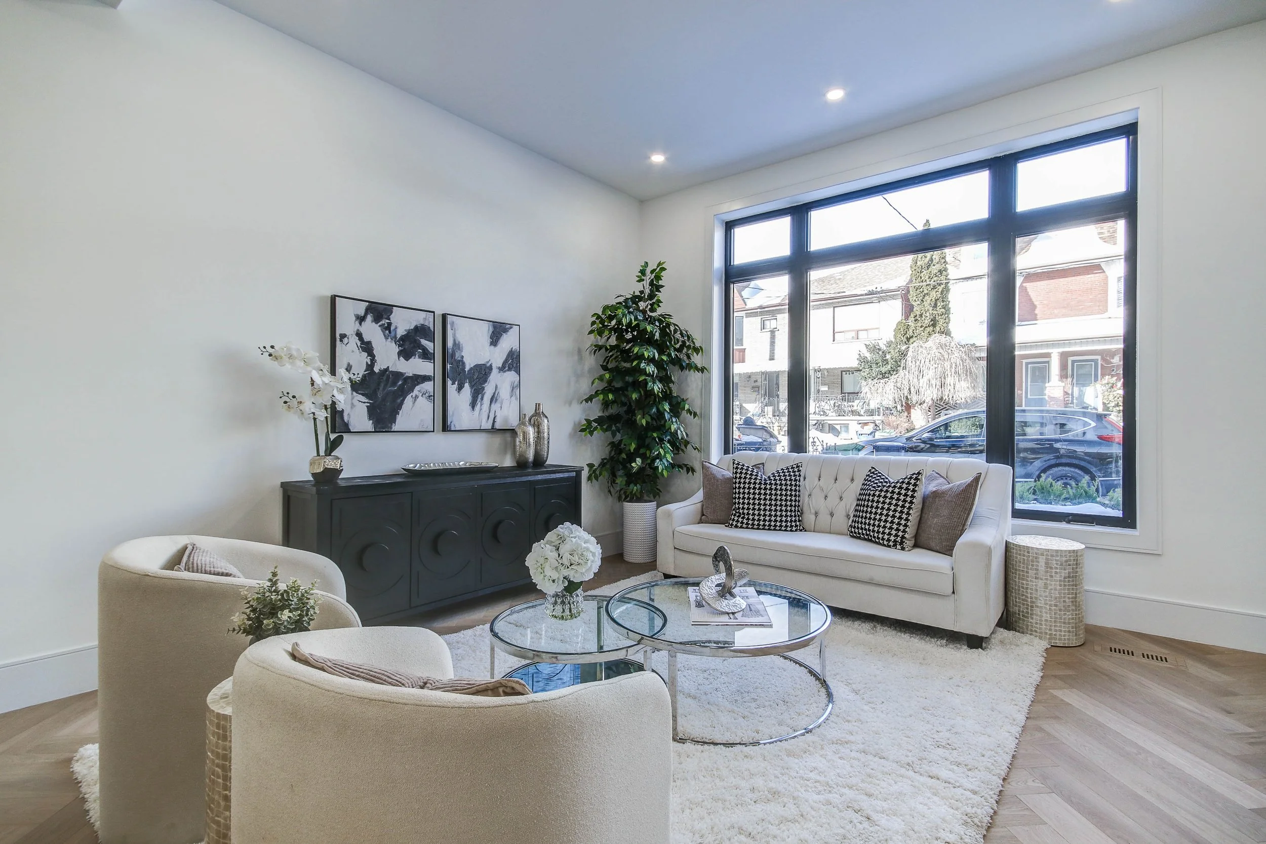 Modern living room with white walls, large window, white tufted sofa with patterned pillows, round glass coffee tables, beige armchairs, black sideboard with white orchids and abstract artwork, green potted plant, and a white shag rug.