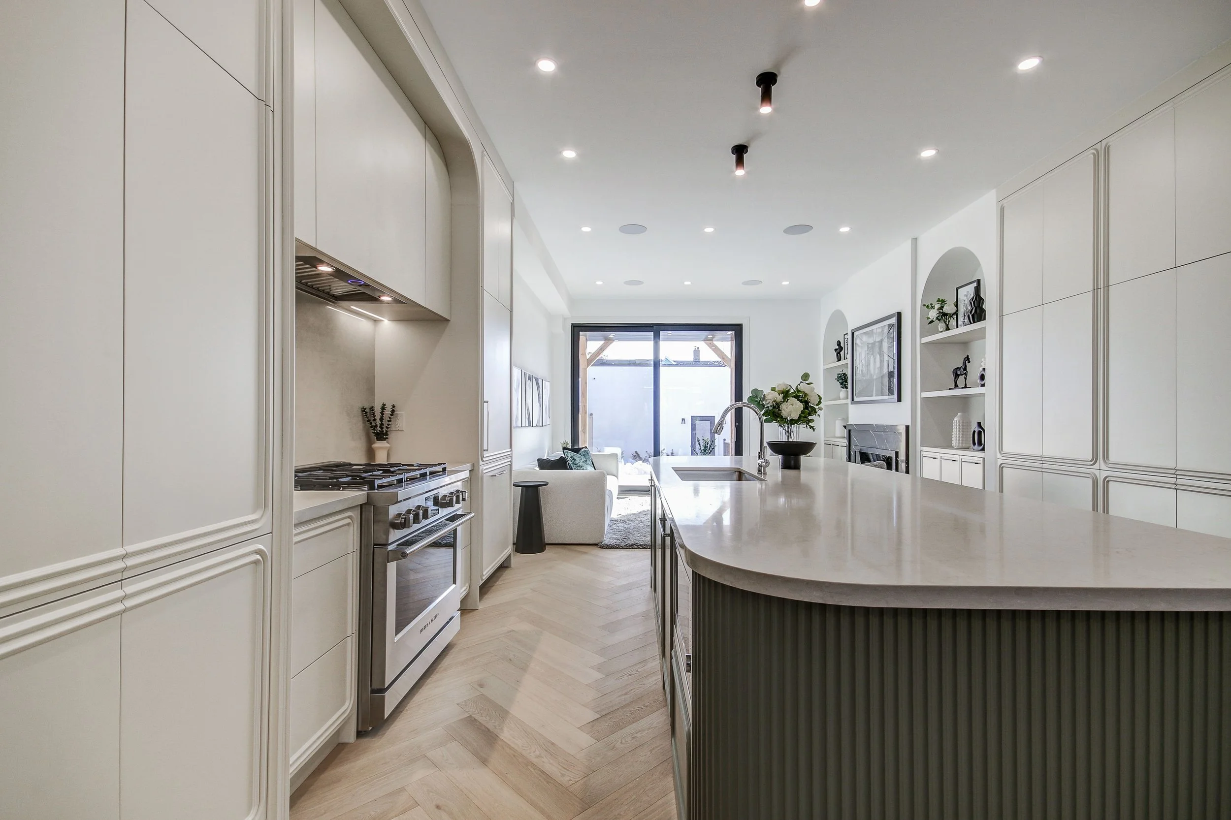 A modern kitchen with white cabinets, a large island with a marble countertop, a gas stove, and a sink. There is a living area visible in the background with a sofa, a large window, and decorative shelves on the wall.
