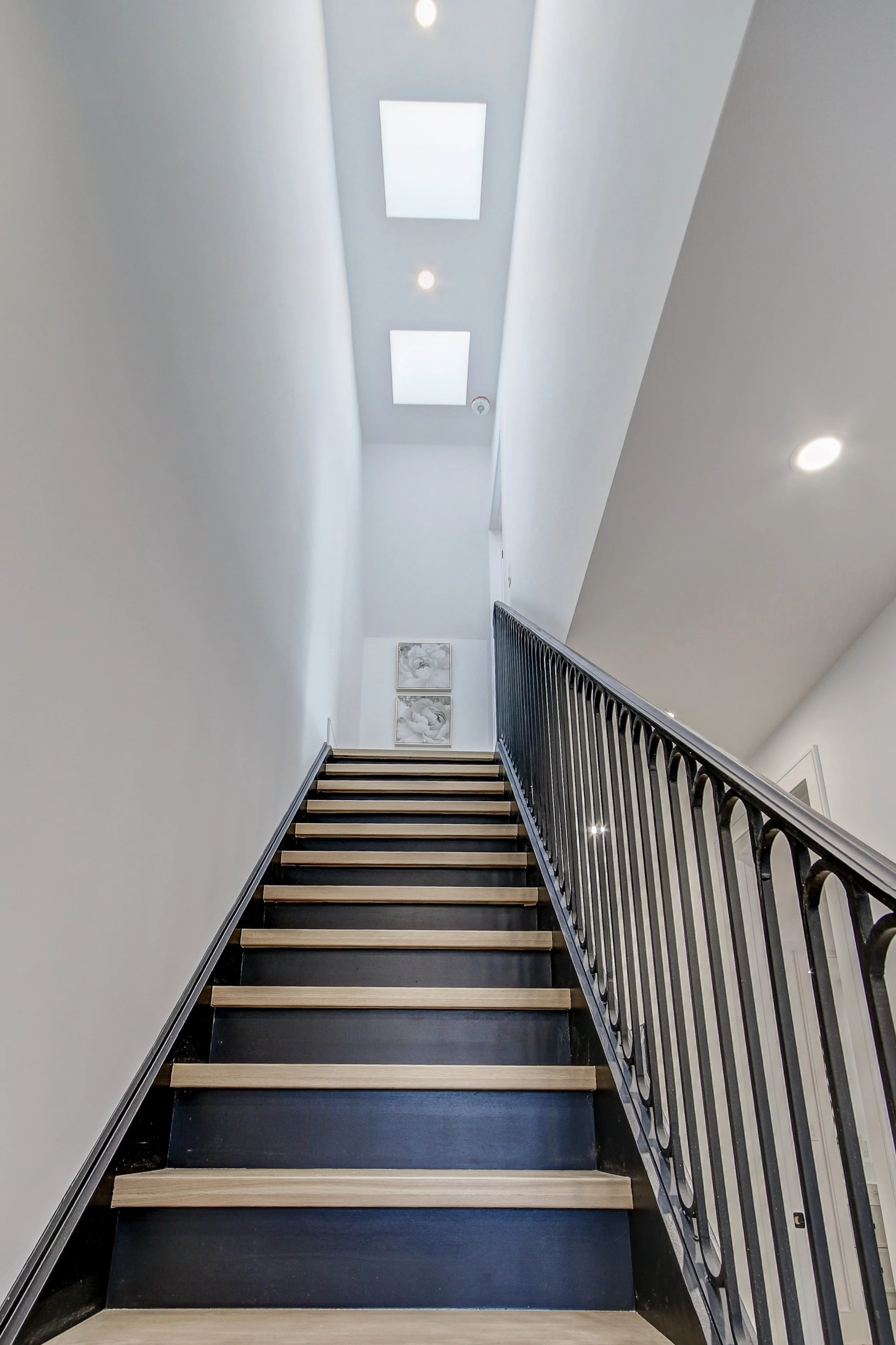 Interior view of a staircase with black steps, wooden treads, and a black and silver metal railing, leading up to a bright white wall with framed artwork, skylights, and ceiling lights.