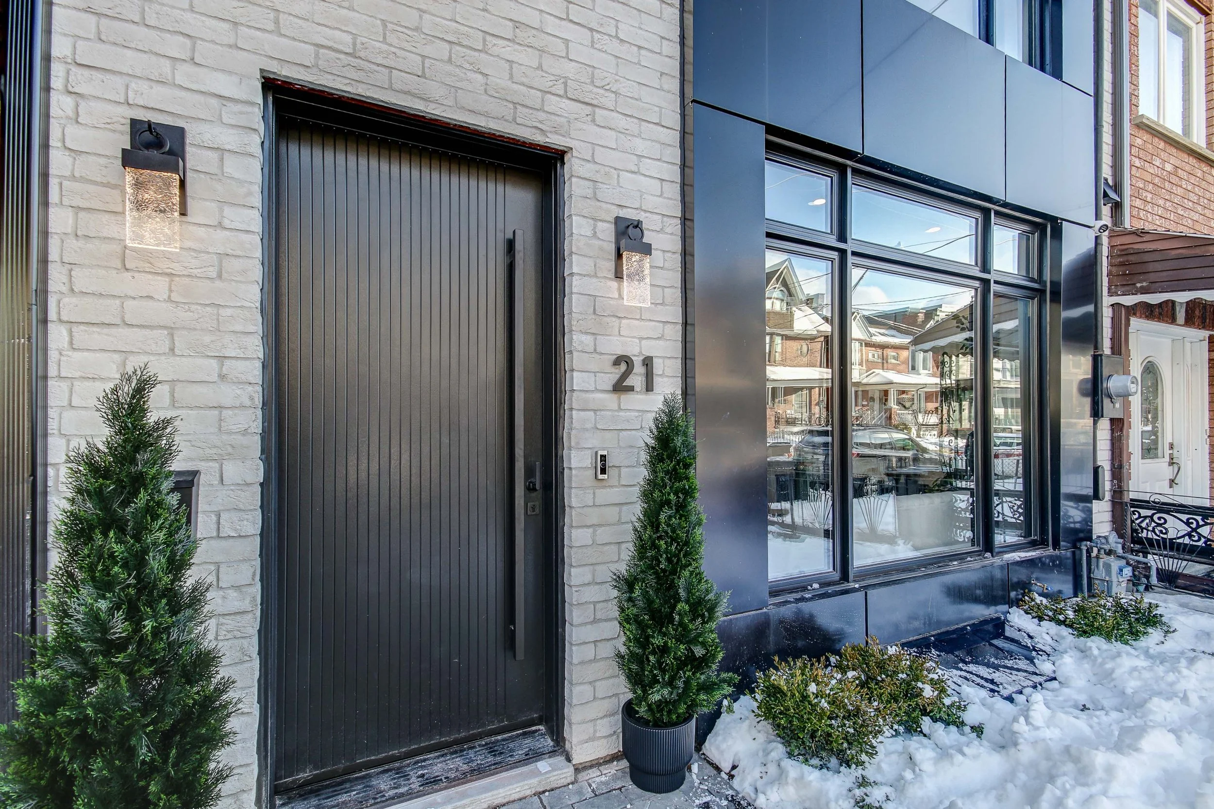 Modern building entrance with black door and glass windows, surrounded by snow and small greenery in pots