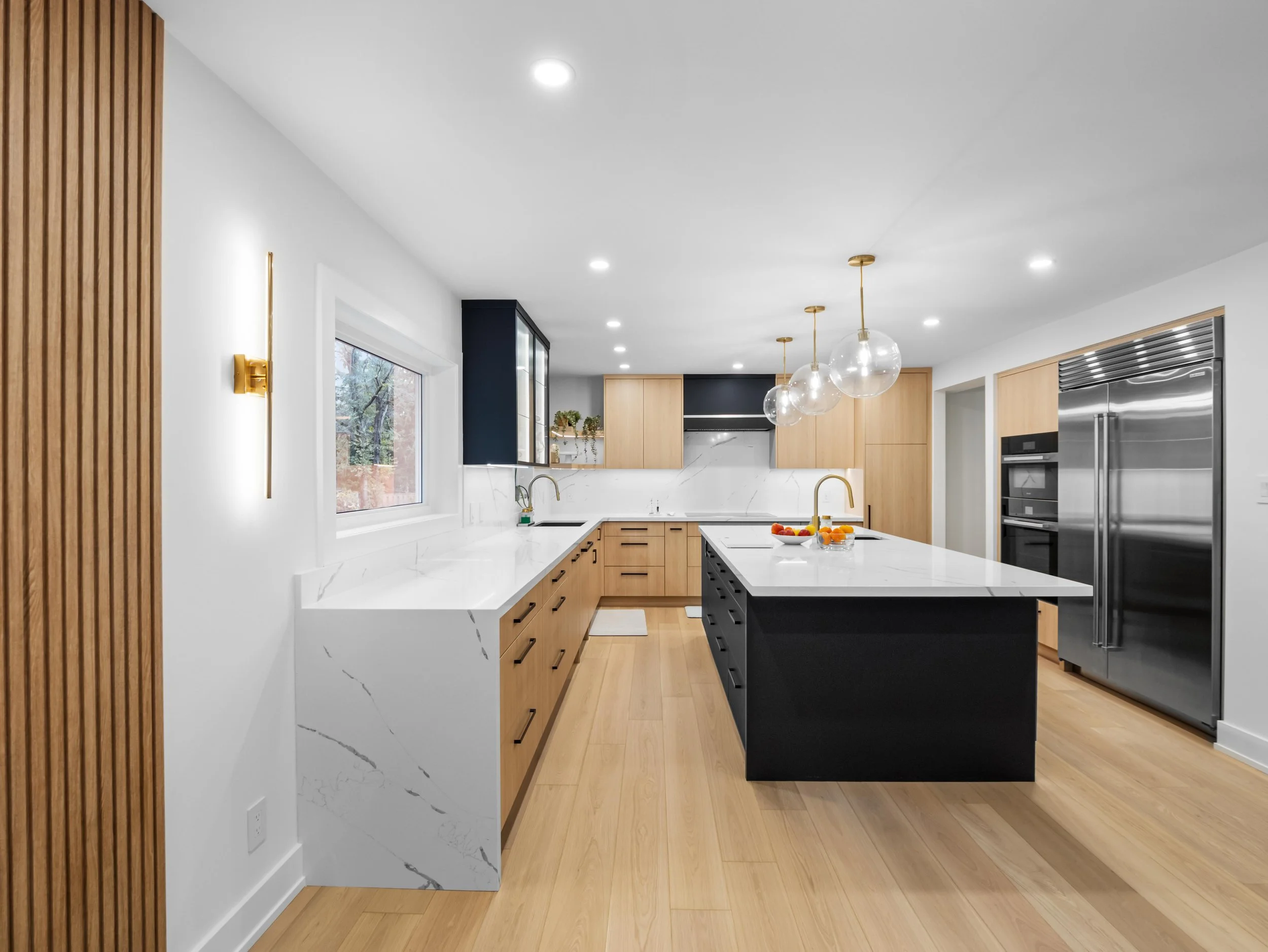 Modern kitchen with white marble countertops, light wood cabinets, a black kitchen island, and three clear pendant lights hanging from the ceiling.