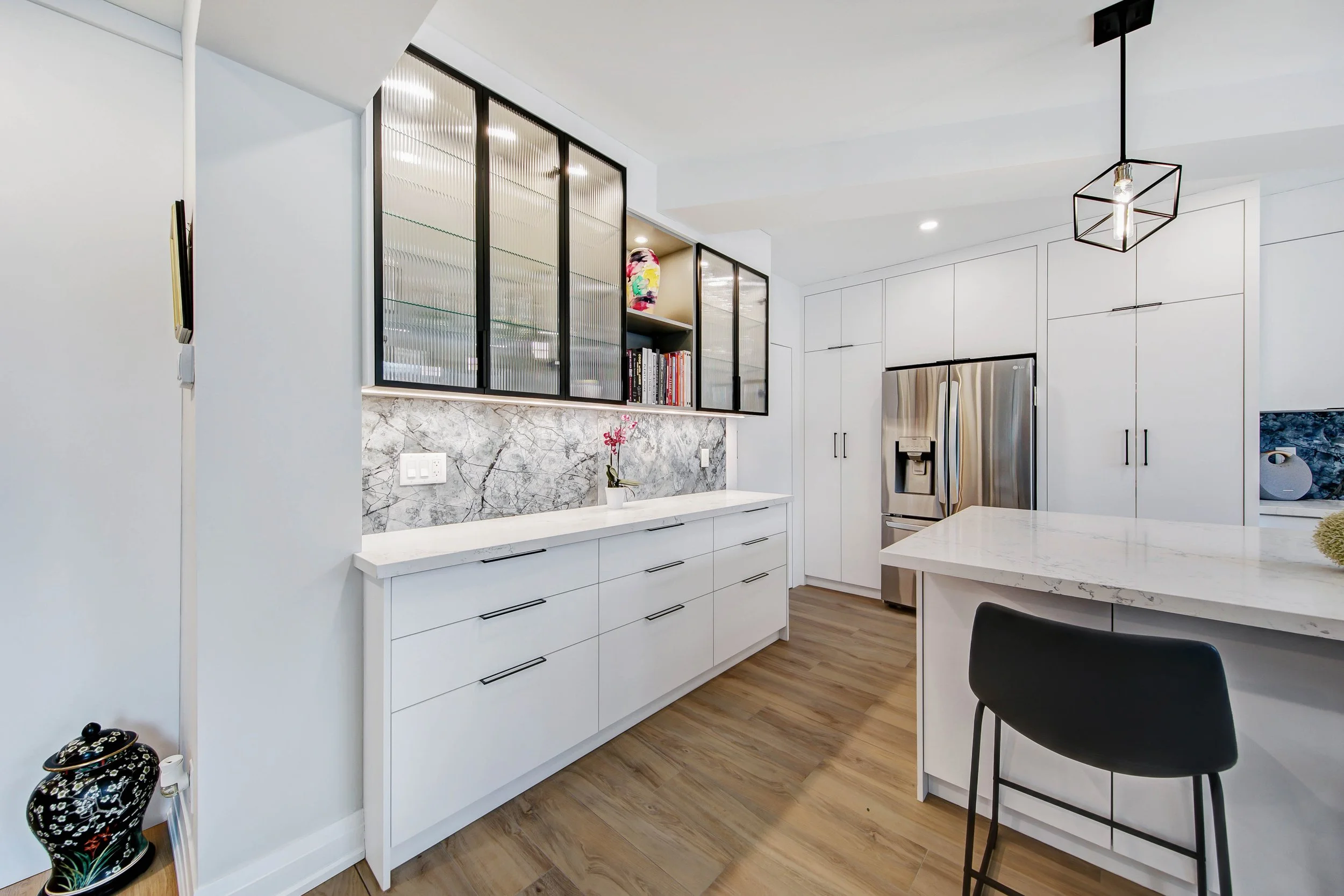 Modern kitchen with white cabinets, marble backsplash, stainless steel refrigerator, black lighting fixture, wooden flooring, and a small table with a black chair.