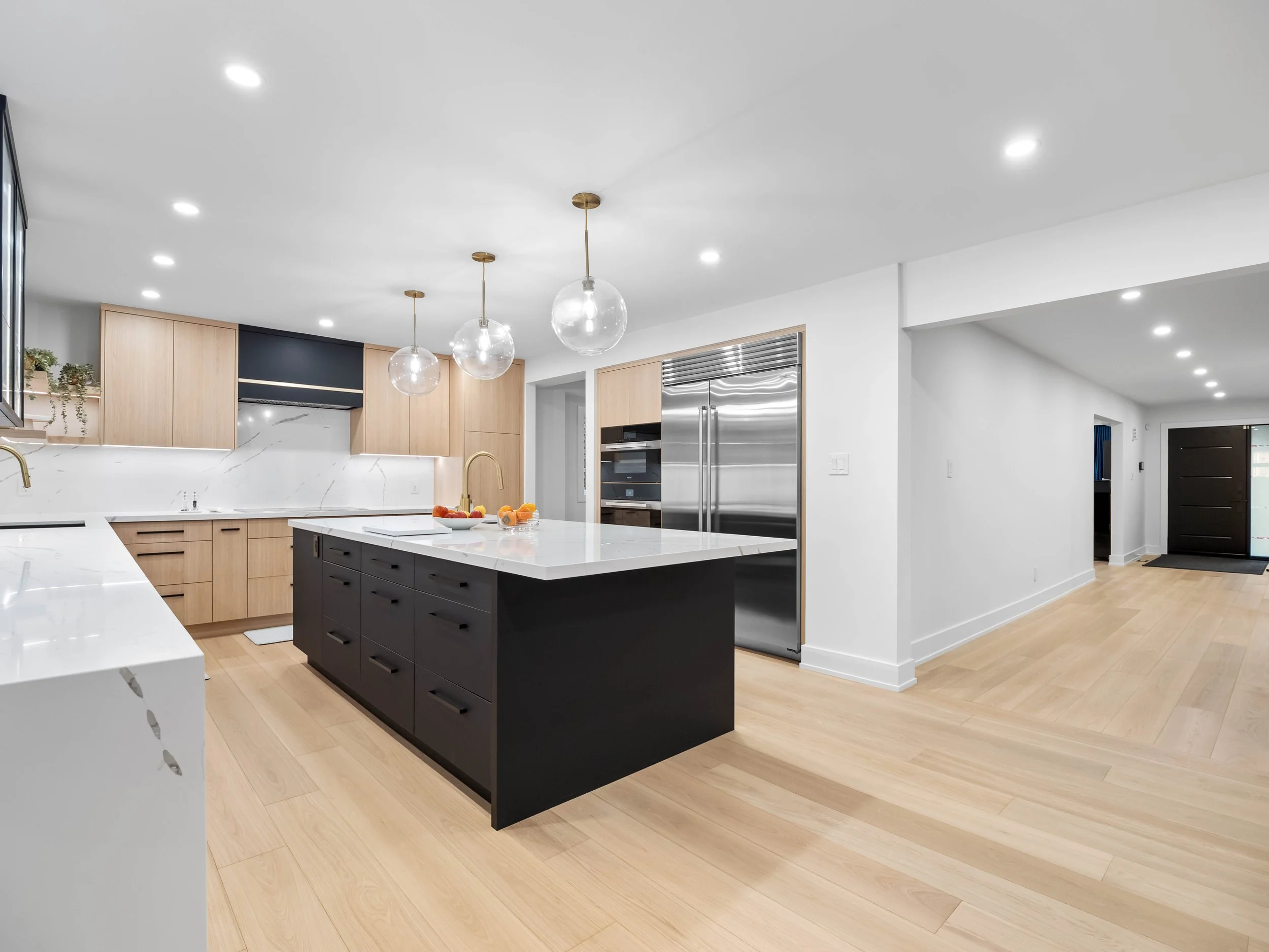 Modern kitchen with white countertops, light wood cabinets, a black kitchen island, stainless steel refrigerator, and hanging light fixtures with globe-shaped glass shades.