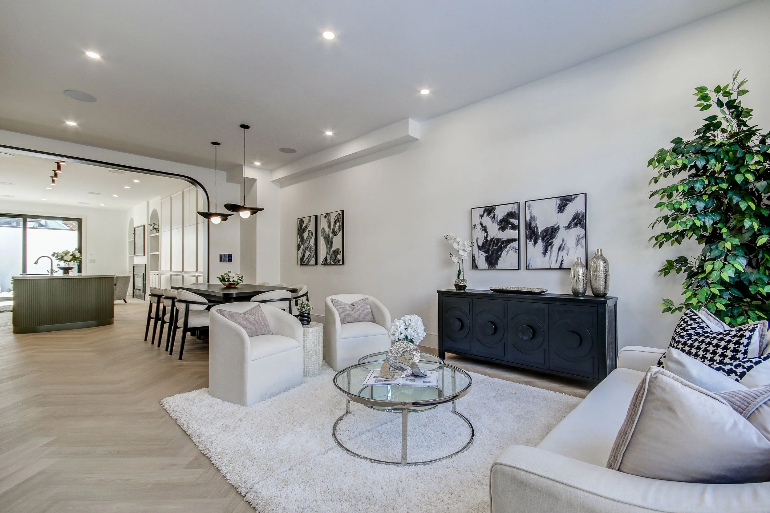 A modern, open-concept living and dining area with white walls, wooden flooring, black and white artwork, and contemporary furniture, including white armchairs, a black sideboard, glass coffee tables, and a large green plant.