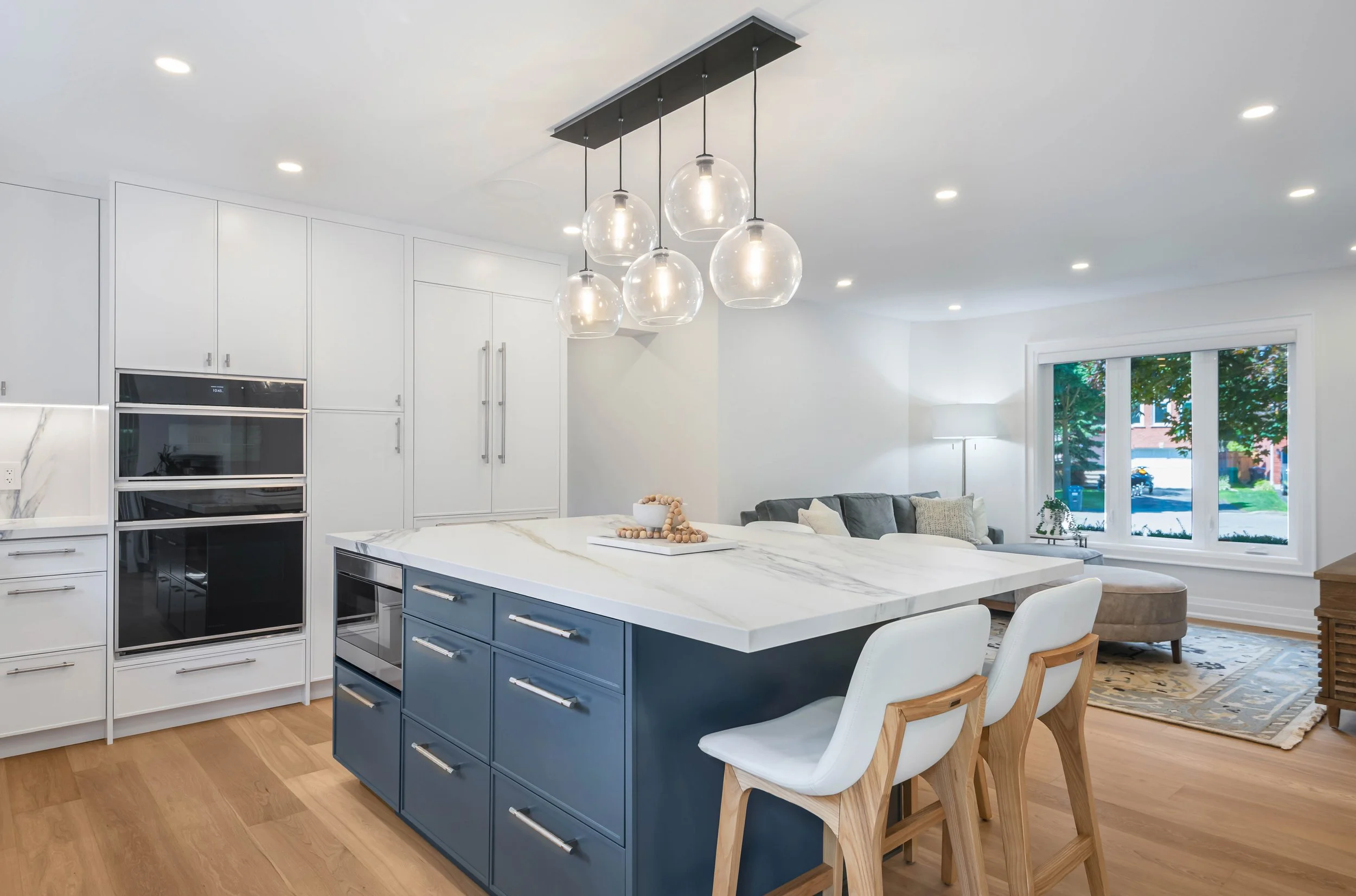 Modern kitchen with white cabinets, a blue island with drawers, a white marble countertop, pendant lights, and a dining area with chairs. Large window with view of trees outside.