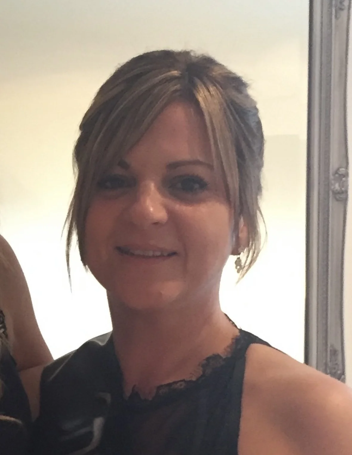 A woman with short, layered blonde hair smiling, wearing earrings and a black lace top, standing indoors near a decorative mirror.