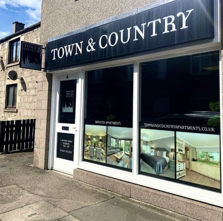 Storefront for Town & Country, a serviced apartments rental business, with large display windows showing interior images of modern living rooms and kitchens, and a sign with contact information.