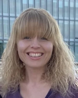 Woman with blonde curly hair smiling in front of a glass building.