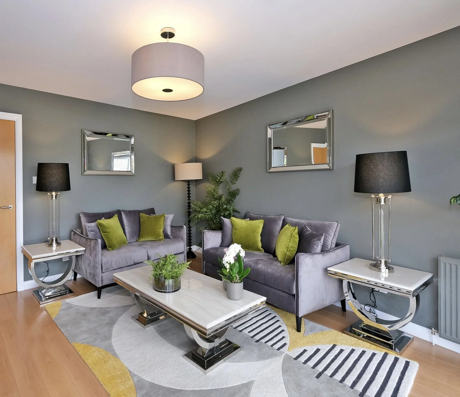 Living room with two gray sofas, black lamps, green pillows, a coffee table with plants, and a patterned rug.