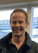 A smiling man with short brown hair and wearing a dark shirt standing indoors near large windows.