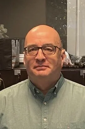 A middle-aged man with glasses and a bald head is wearing a light green button-up shirt in an indoor setting.
