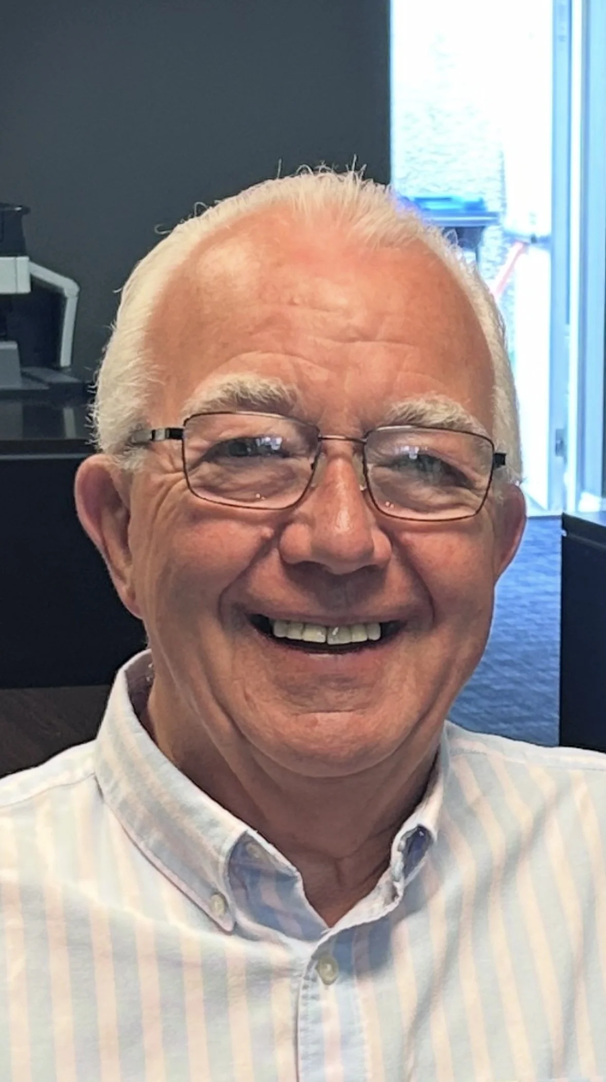 Smiling elderly man with white hair, glasses, and a striped dress shirt in office setting.