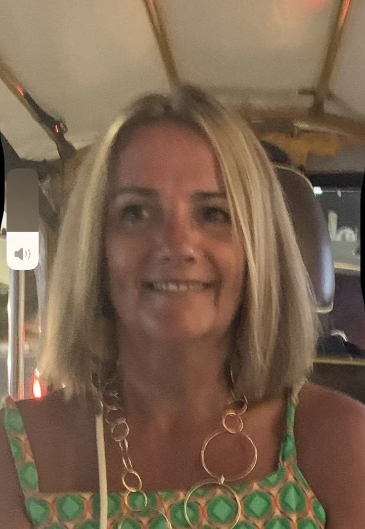 A woman with shoulder-length blonde hair smiling, wearing a green patterned dress and chunky gold necklace, seated inside a vehicle at night.