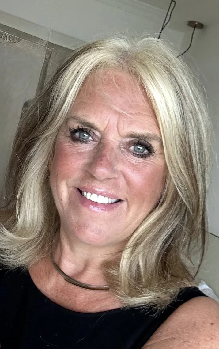 Close-up of a smiling woman with shoulder-length blonde hair, light skin, wearing a black top and a simple necklace.