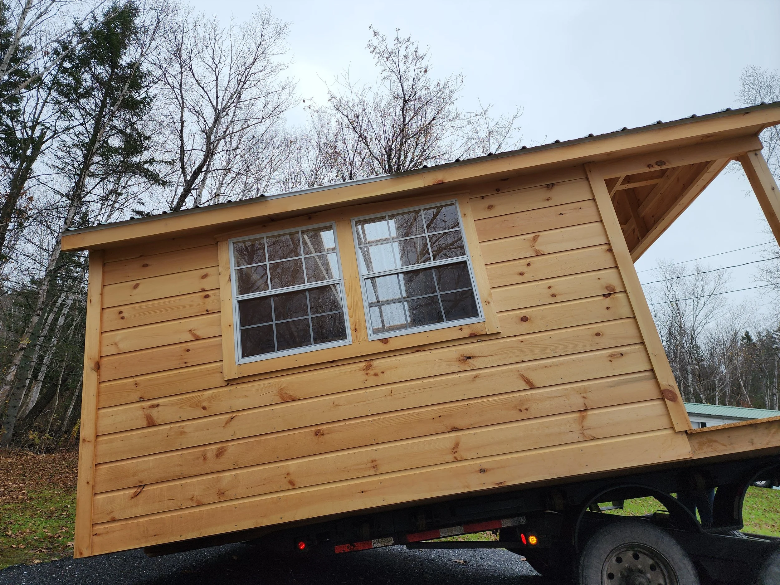 A small wooden house with horizontal pine boards, double-hung windows, set on a flatbed trailer, in a wooded area with leafless trees in the background.