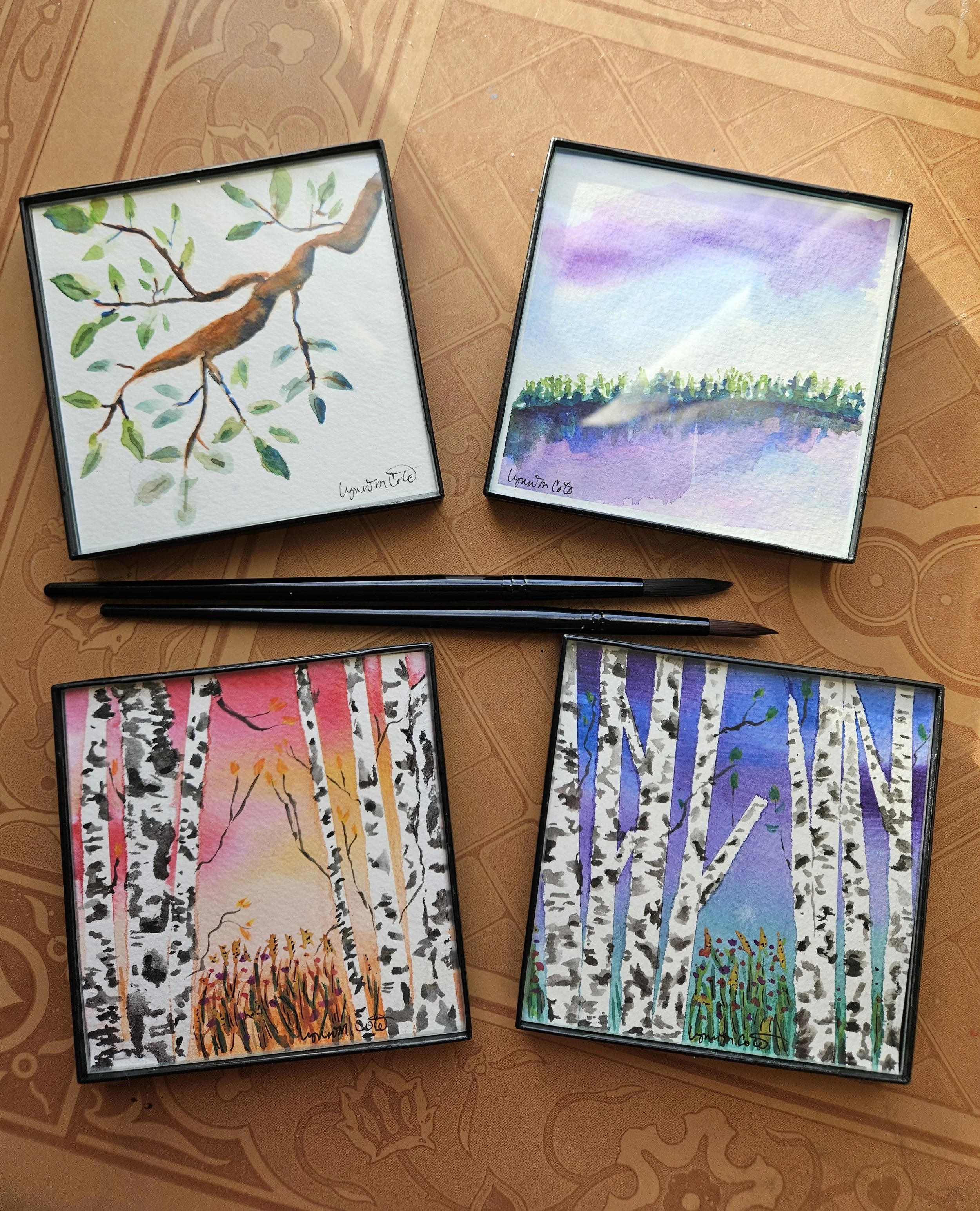 Four small watercolor landscape paintings in black frames, with two paintbrushes in the middle, laid out on a orange patterned tablecloth. The paintings depict trees and nature scenes in various colors including greens, purples, pinks, and oranges.