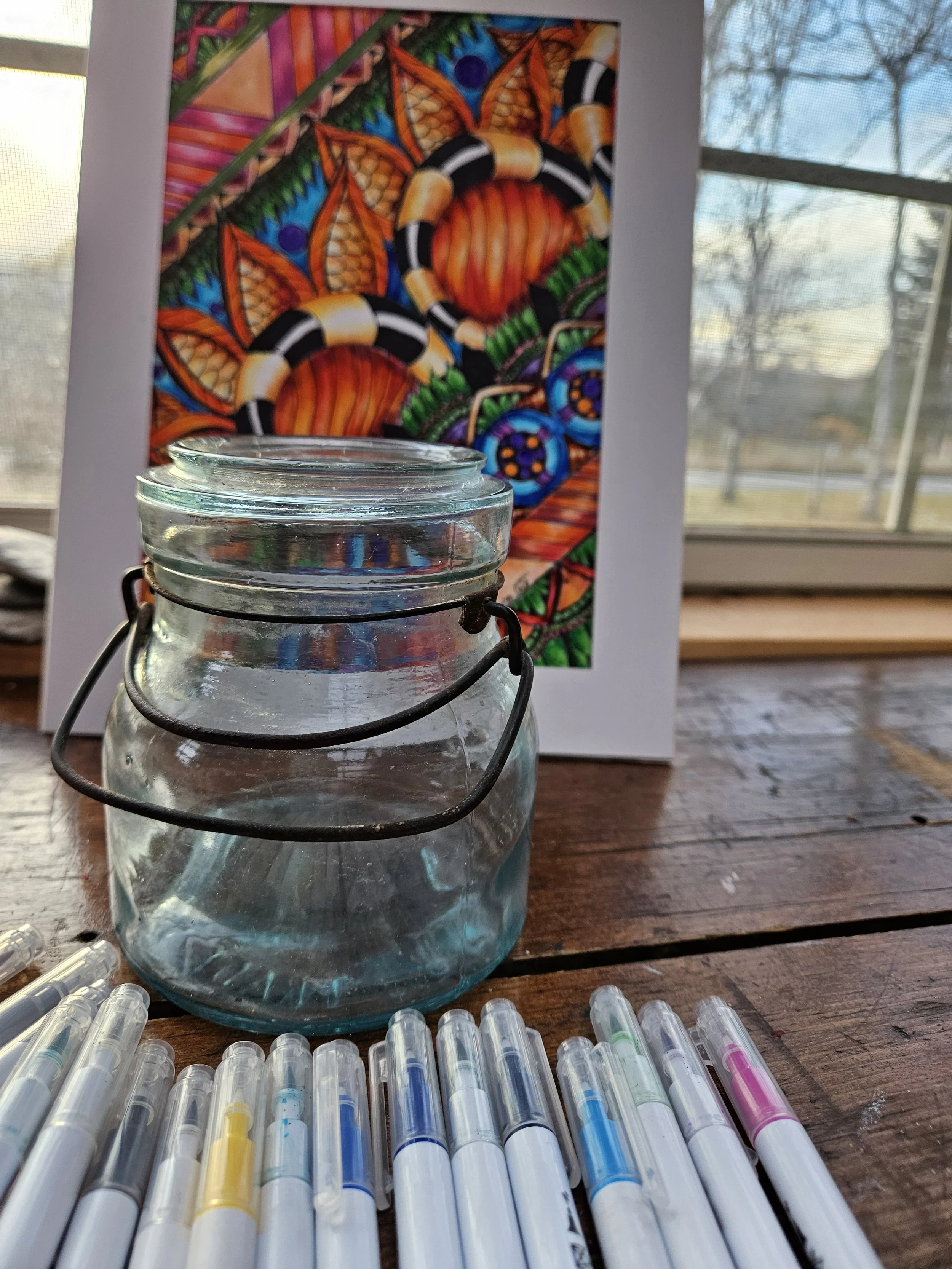 A glass jar with a metal handle in the foreground, surrounded by colorful markers, in front of a vibrant, abstract, colorful painting on a table. The background includes a window with a view of trees outside.