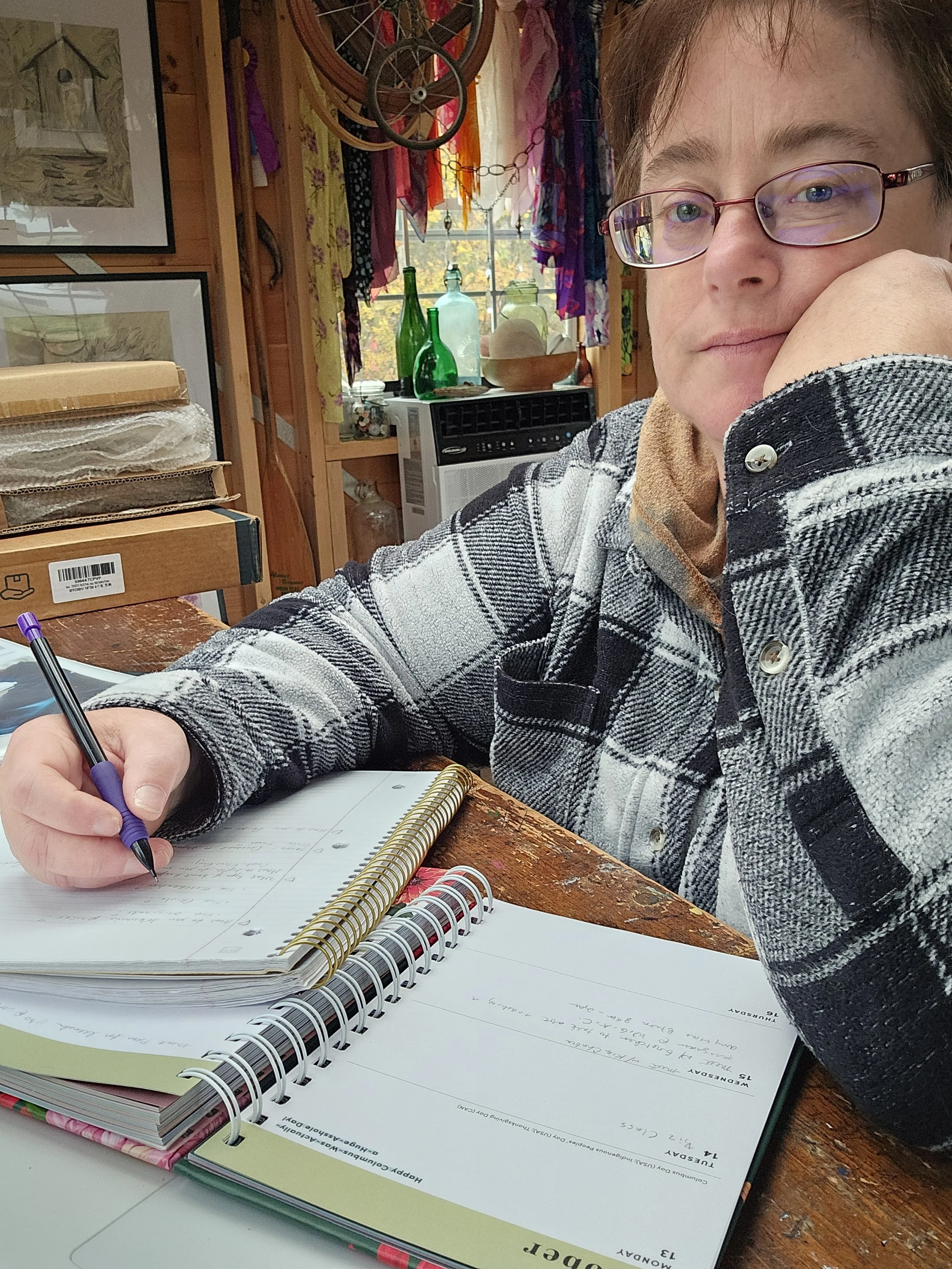 A person with short hair and glasses sitting at a wooden table, writing in a notebook. The background shows a room with colorful fabric hanging, glass bottles on a shelf, and framed artwork.