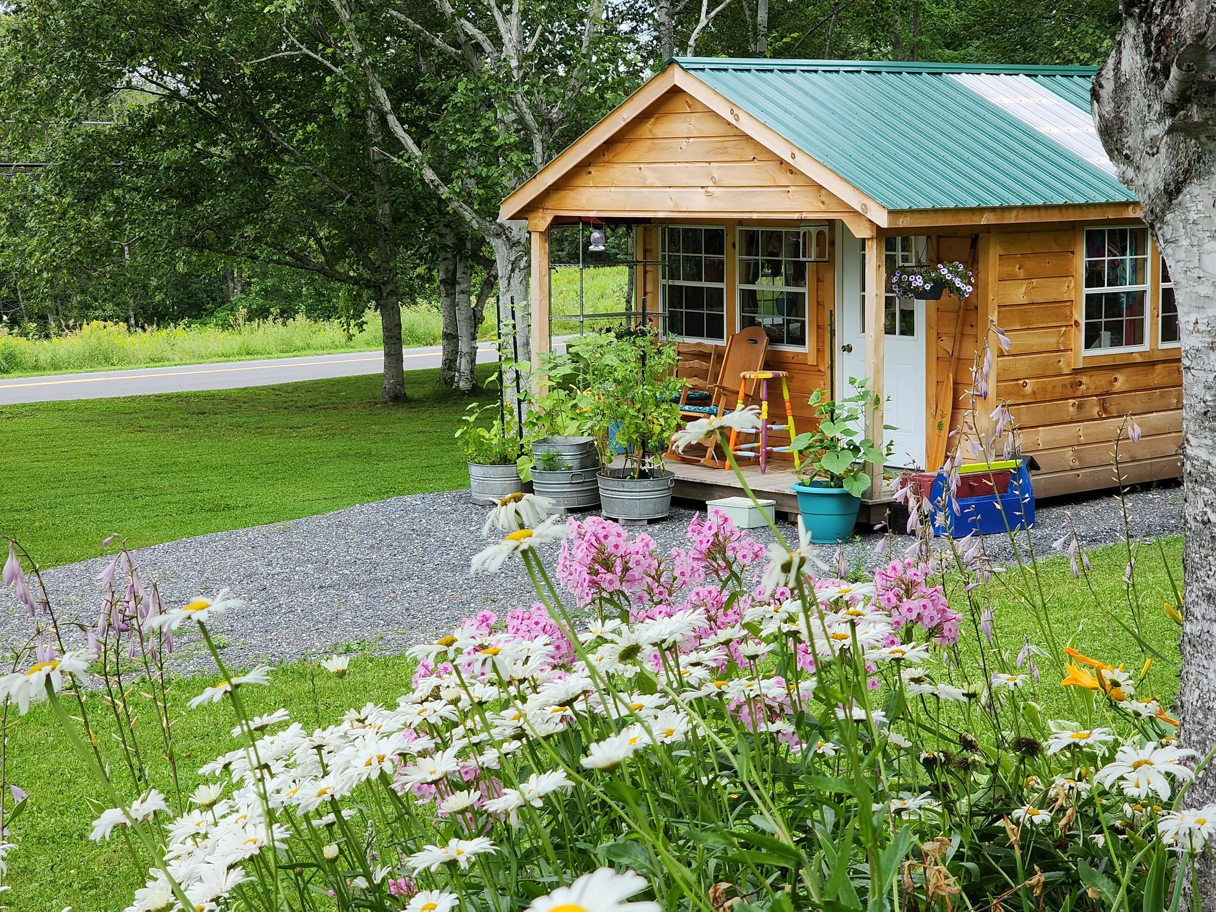 A wooden garden shed with a green metal roof, surrounded by colorful flowers, potted plants, and a lawn. A gravel path leads to the shed, and there are trees in the background.