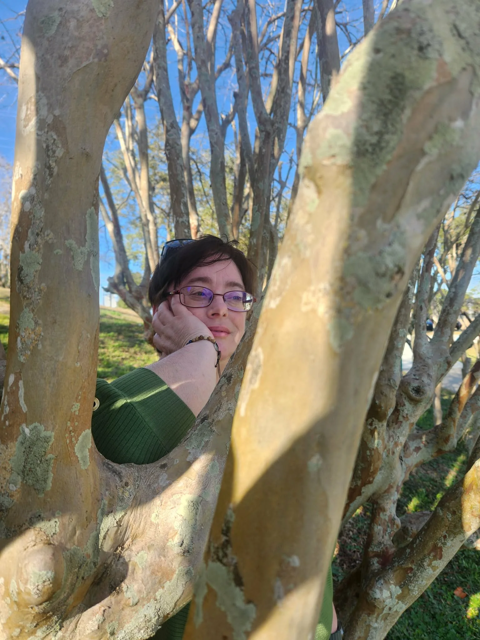 A woman with short dark hair and glasses resting her head on her hand, looking through a tree's branches with peeling bark and green moss in a park on a sunny day.