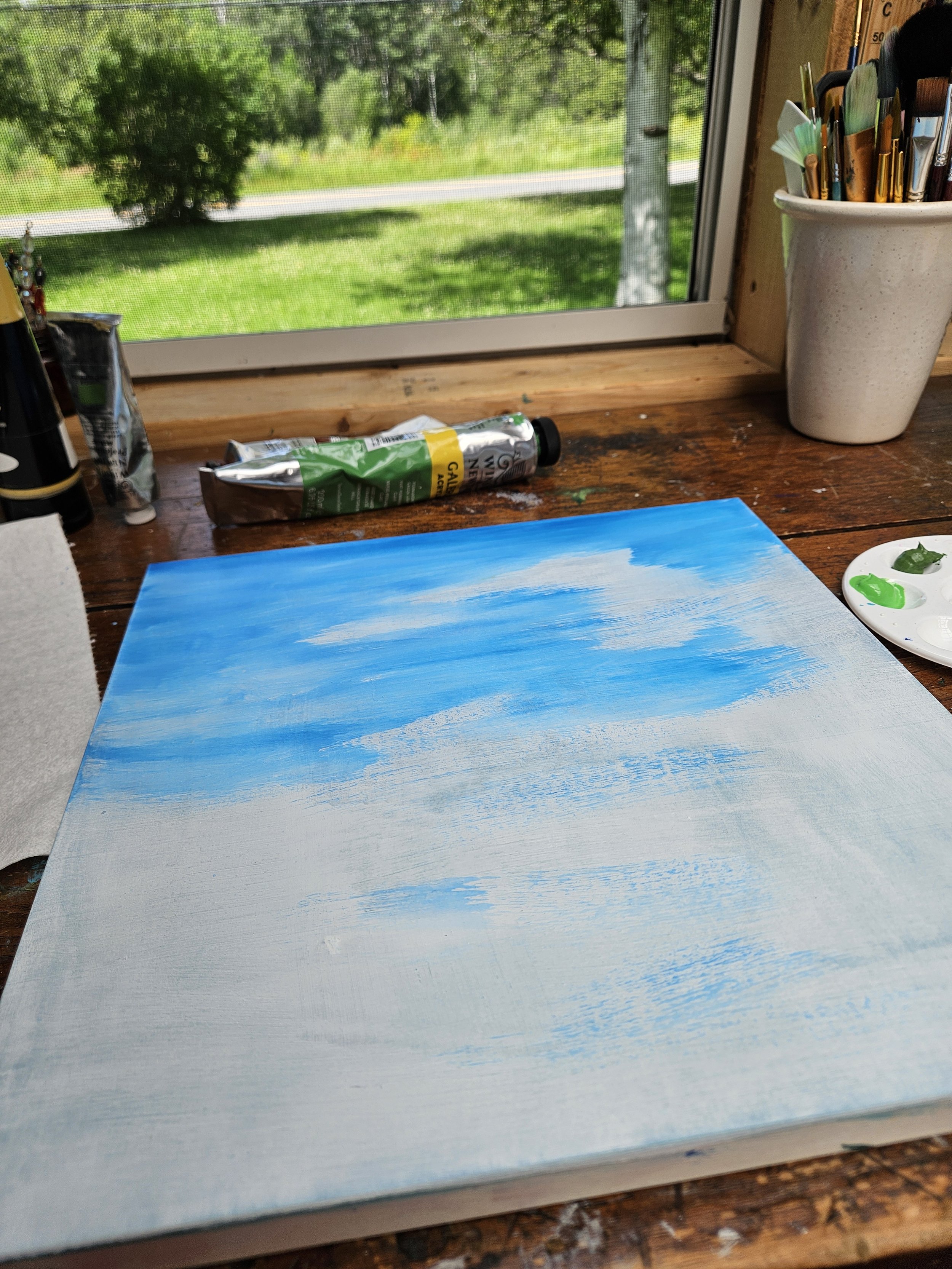 A canvas with a partly painted sky scene in progress, featuring blue and white clouds, on a wooden table near a window with a view of green trees and grass outside. Art supplies like paint tubes, brushes, and a palette are nearby.
