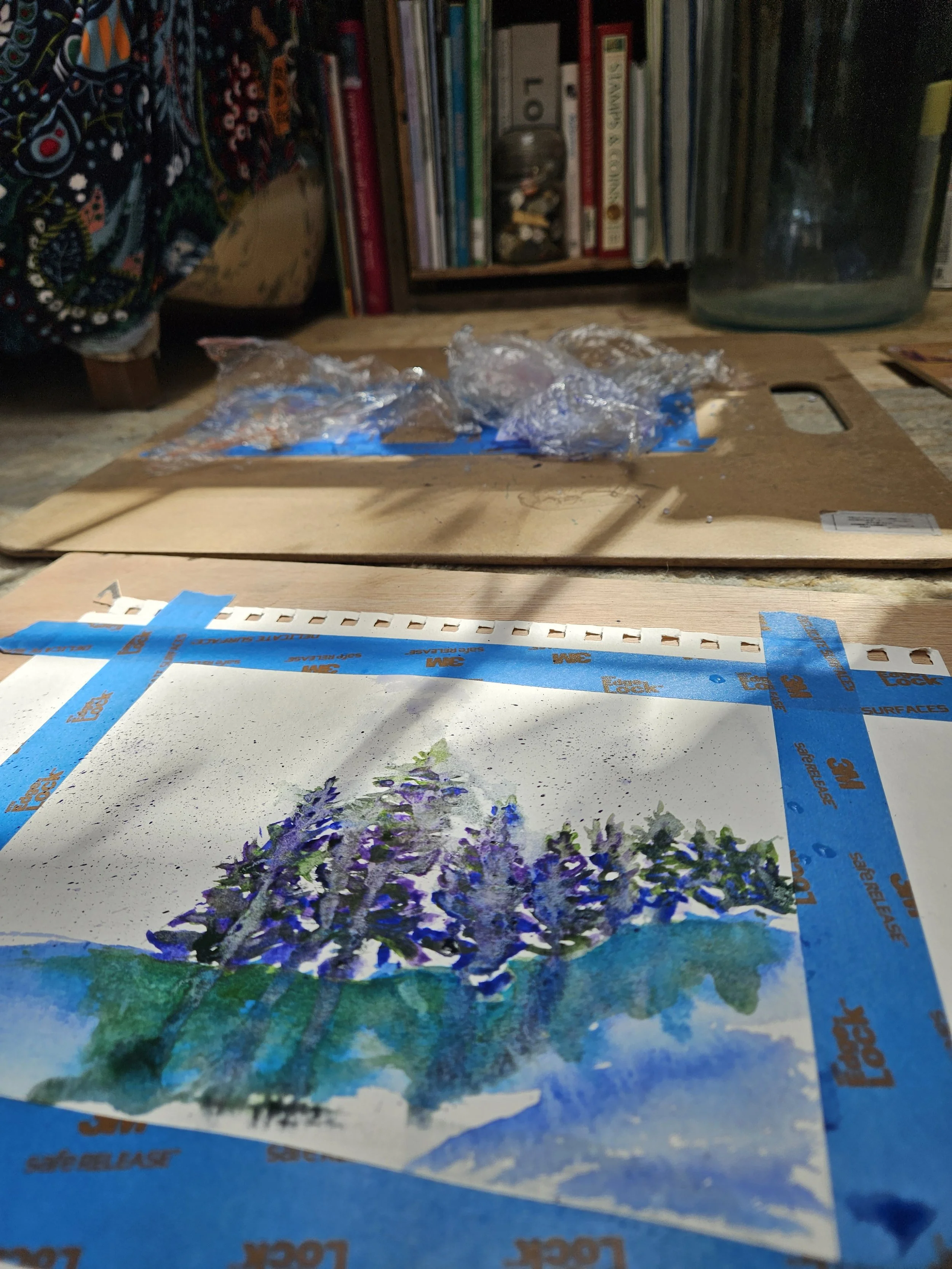 A watercolor painting of a purple flowering tree on paper taped down with blue painter's tape, with a background of a bookshelf and art supplies.