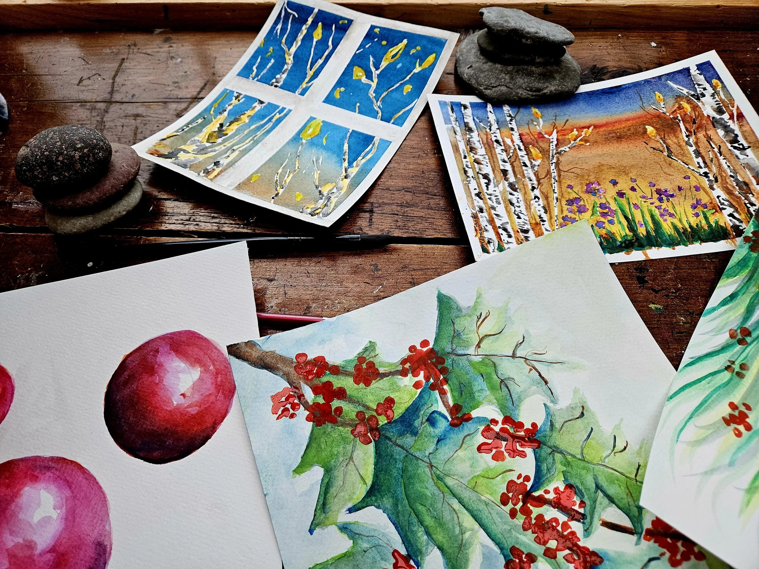 Watercolor paintings of a sunset, birch trees, apples, and holly leaves with red berries are spread on a wooden surface, along with small stacks of natural stones.