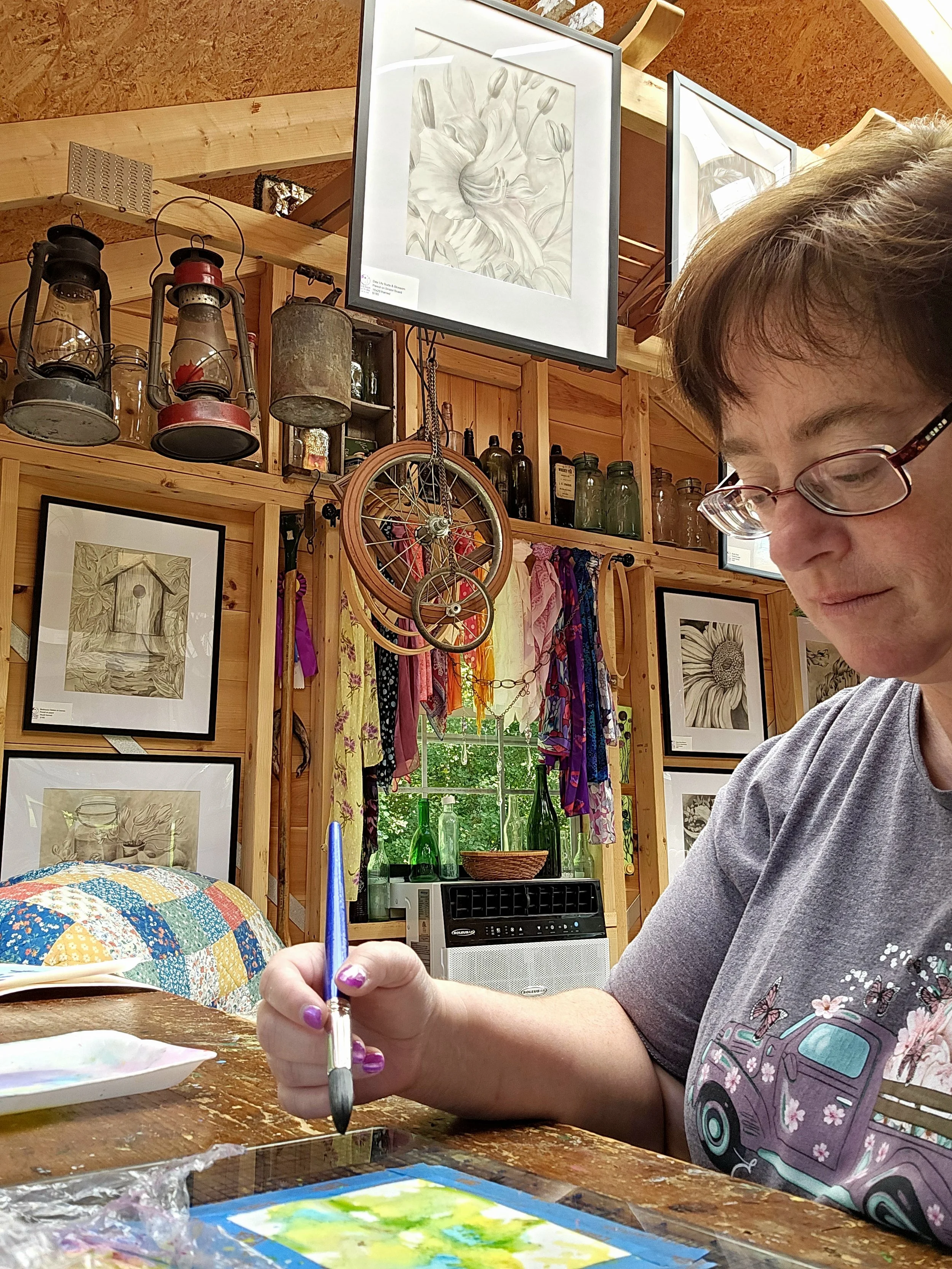 A woman with glasses painting watercolor art at a wooden table in a cozy, rustic room decorated with framed botanical prints, vintage lanterns, a bicycle wheel, glass bottles, and colorful fabric hanging by a window.