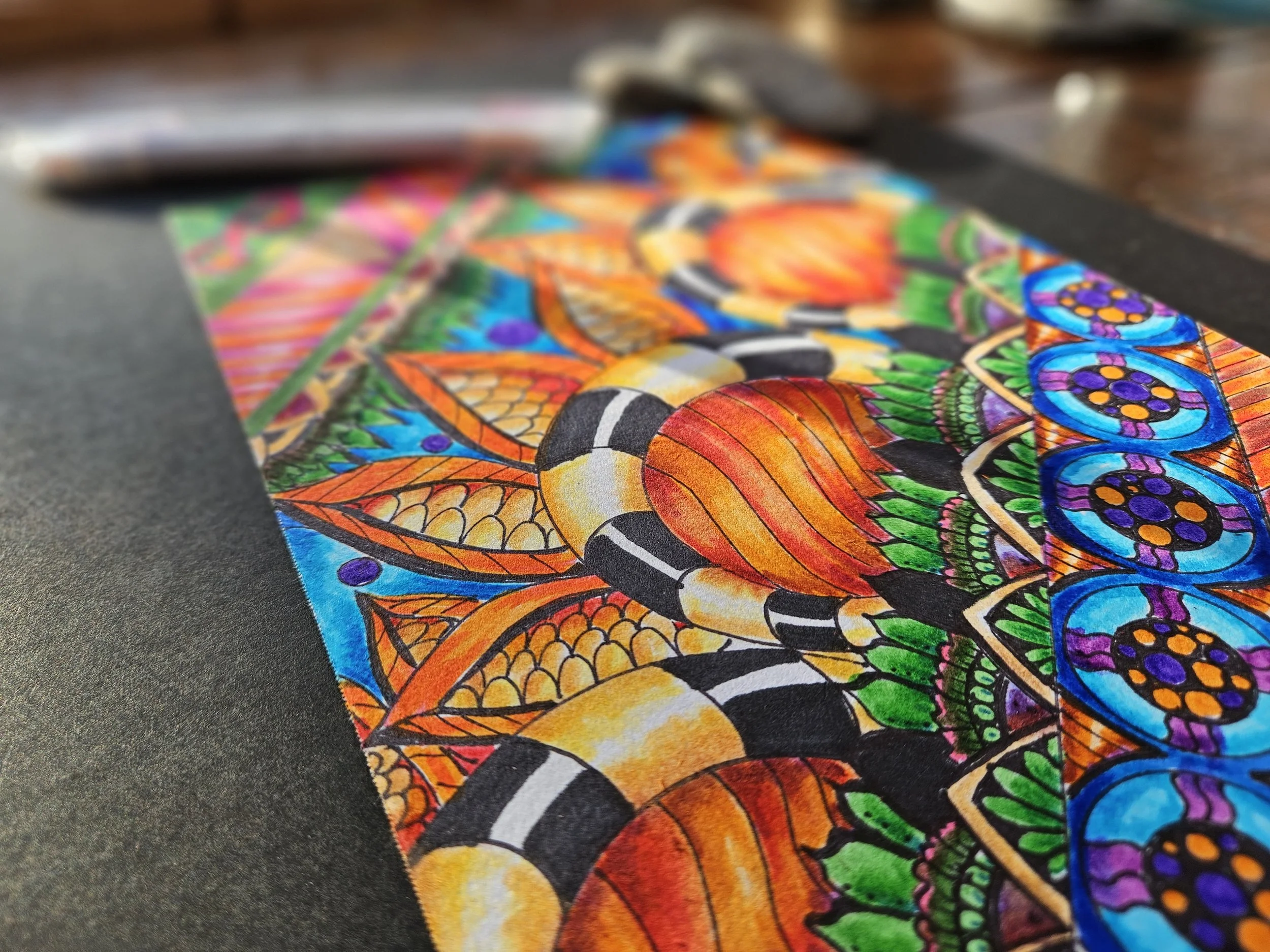 Close-up of a colorful, detailed mandala-inspired drawing on paper, featuring orange, yellow, blue, green, purple, and black intricate patterns.
