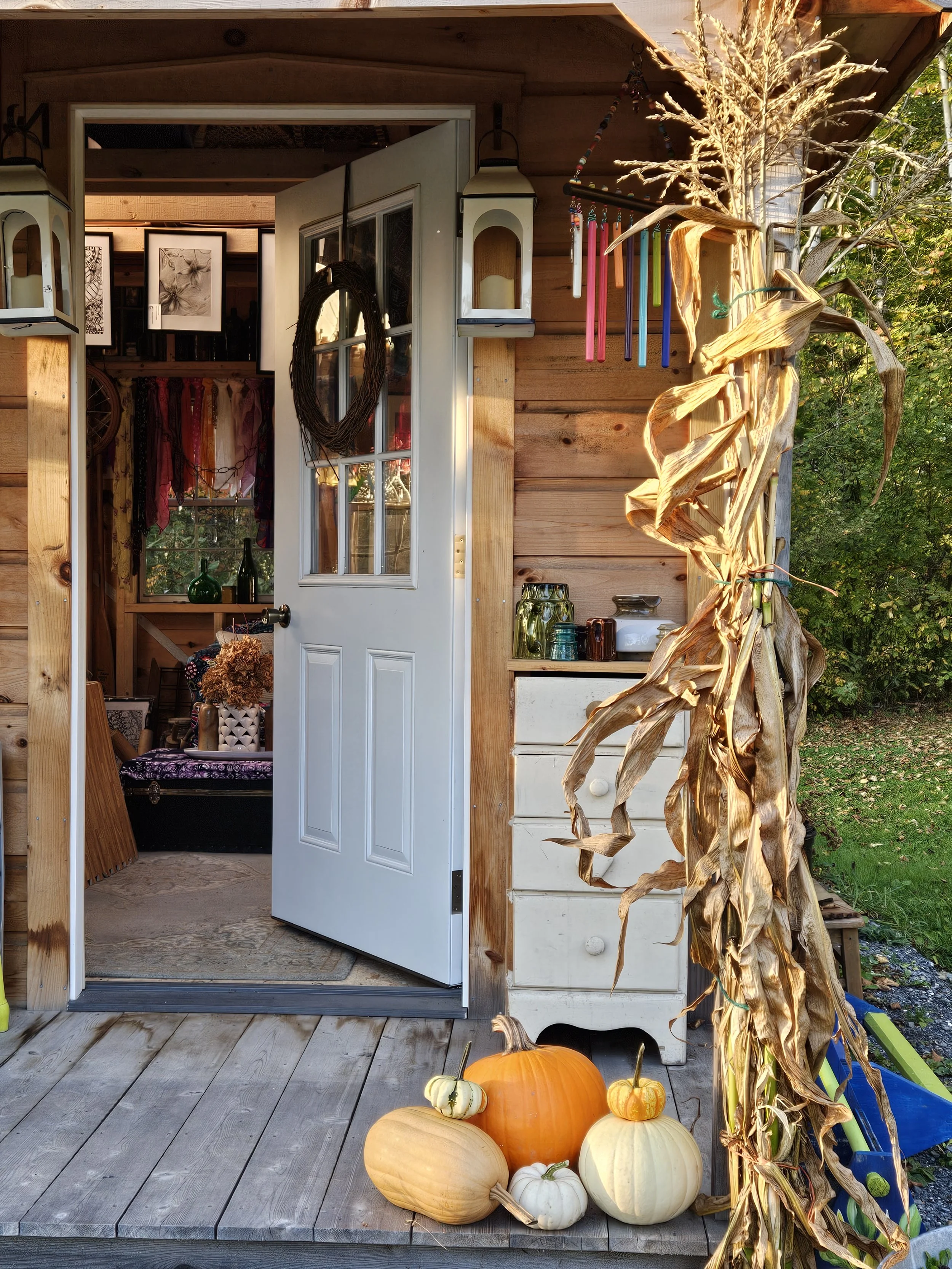 An outdoor porch scene with a white door open to a cozy interior. On the porch steps, there are various pumpkins and gourds, and corn stalks are tied to the side. Decorative lanterns and colorful sticks hang from the overhang, with trees and greenery in the background.