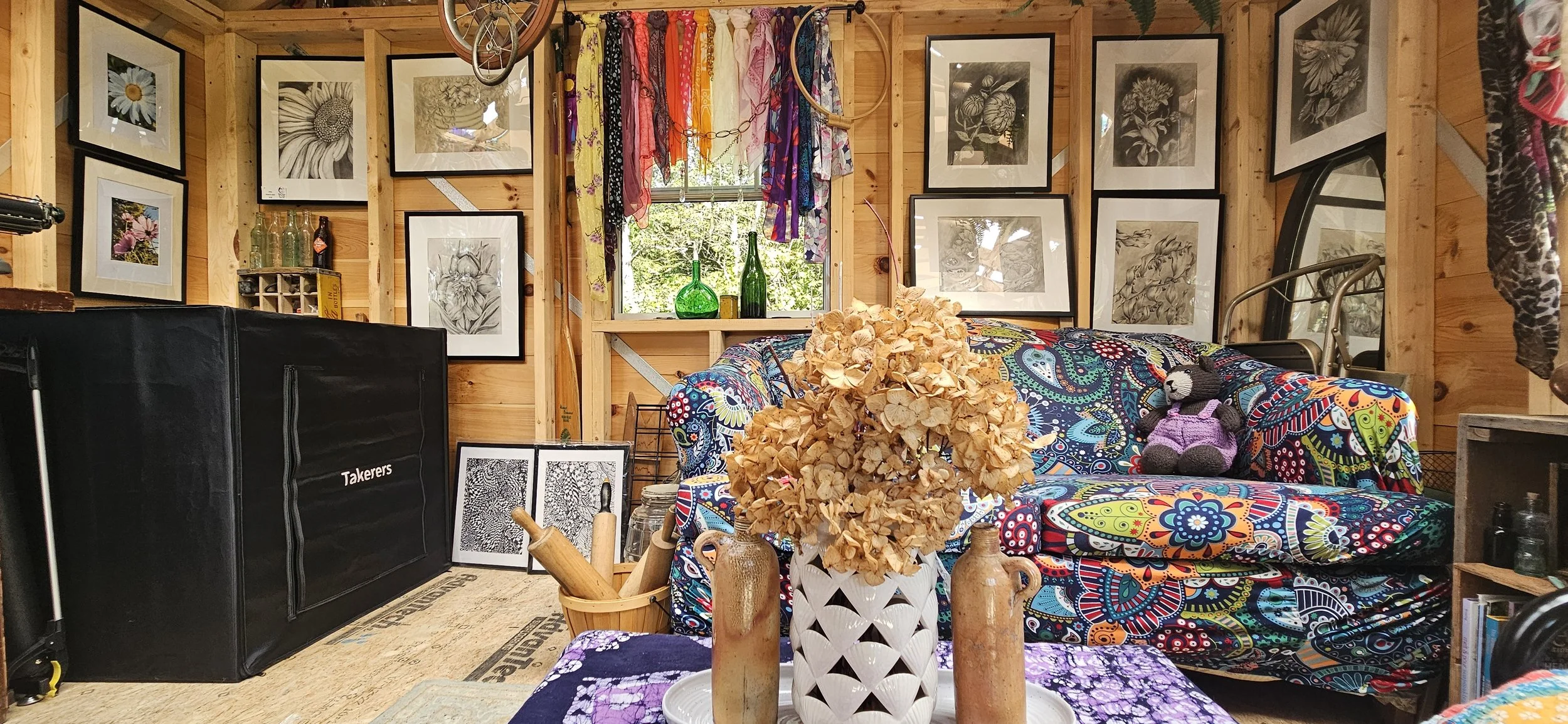 A cozy living room with wooden walls and framed black-and-white and color photographs of flowers hanging on the walls. A window with colorful scarves hanging as curtains lets in natural light, revealing green foliage outside. There is a colorful patterned couch with a stuffed bear, a vase with dried flowers, and decorative bottles on the windowsill. A table with books and pottery is in front of the couch, and a black cabinet labeled "Taxerers" is on the side. Various decorative items, including framed artwork and bottles, are arranged throughout the room.