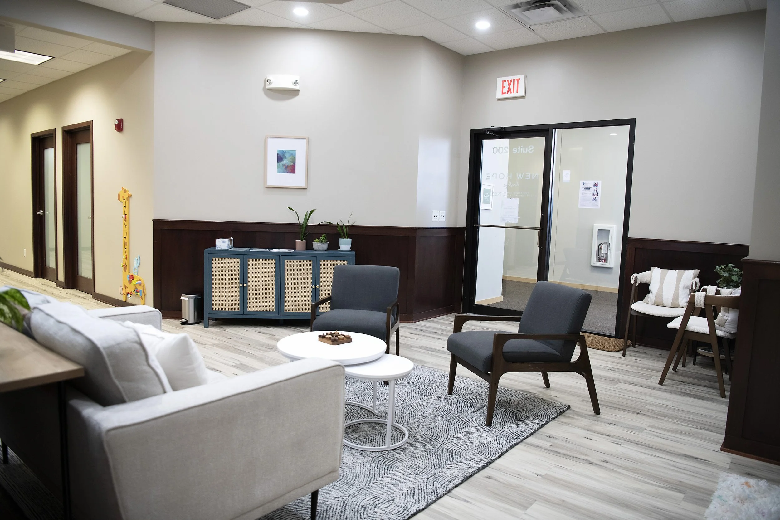 A modern waiting room with beige sofa, gray armchairs, coffee tables, potted plants, framed artwork, and a door leading outside. There is a blue cabinet and children's giraffe decoration on the wall.