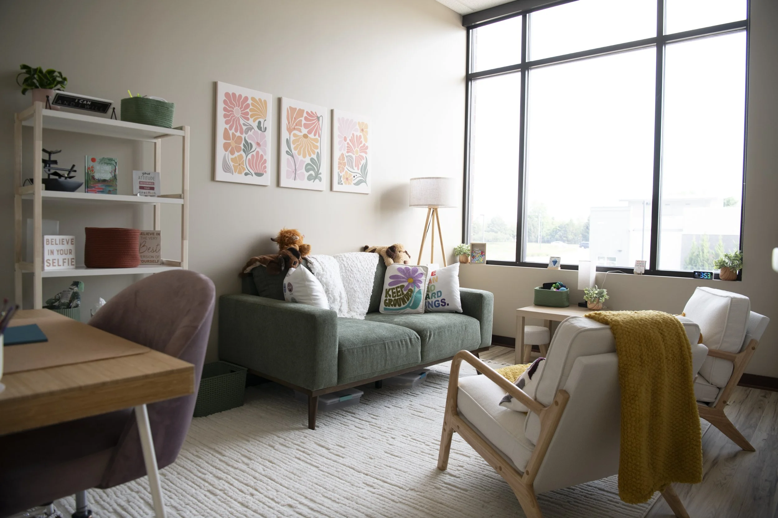 Living room with green couch, two white armchairs, large window, wall art, bookshelf, lamp, decorative pillows, and various plants