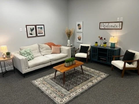 A cozy waiting room with a white sofa, decorative pillows, a wooden coffee table on a patterned rug, a black and white armchair, a black sideboard with a lamp and plants, and wall art including framed pictures and a welcome sign.