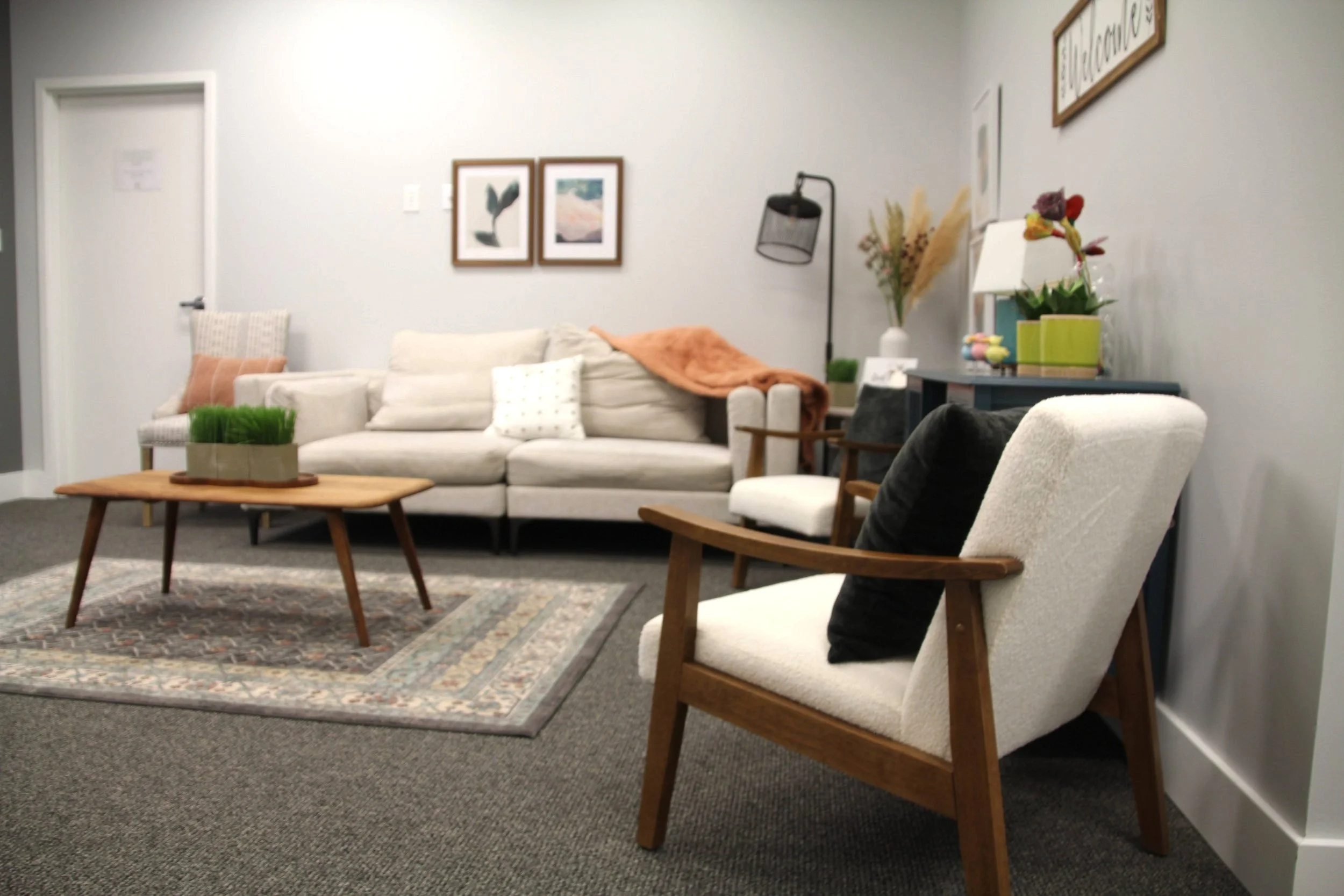 Cozy waiting room with beige sofa, white armchair, wooden coffee table, and wall art, featuring decorative pillows and plants.