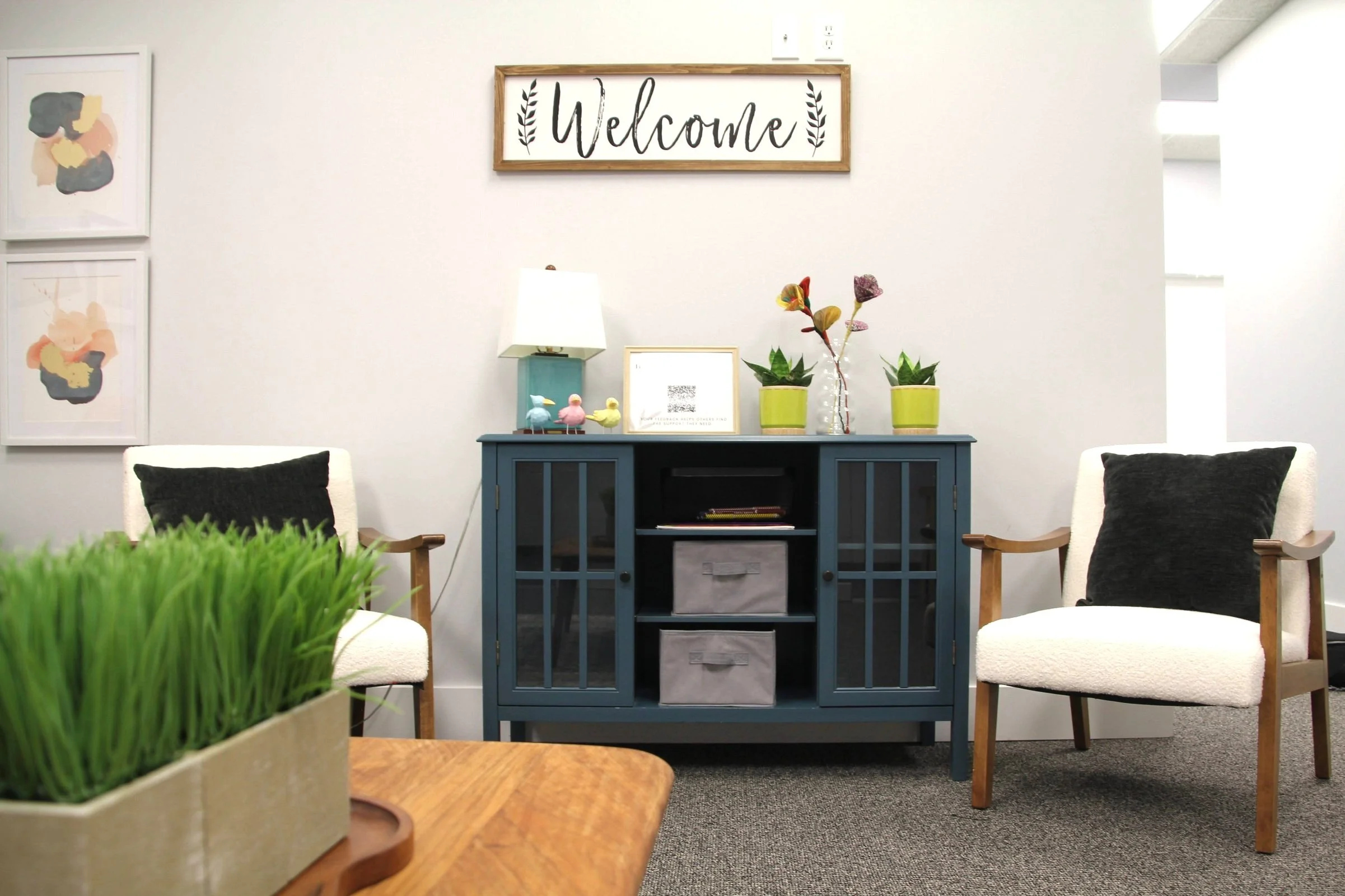 Cozy waiting area with two white armchairs with black pillows, a blue cabinet with potted plants and decorative items, a framed welcome sign on the wall, and a wooden table with a green plant in the foreground.