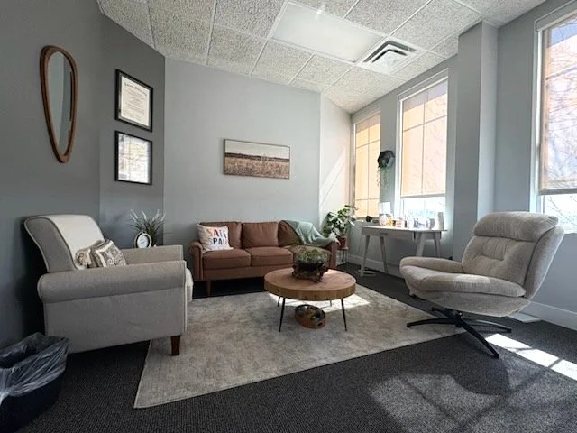 A modern therapy office with a gray wall, framed certificates, a brown sofa, two beige armchairs, a wooden coffee table, a light gray rug, and large windows letting in natural light.