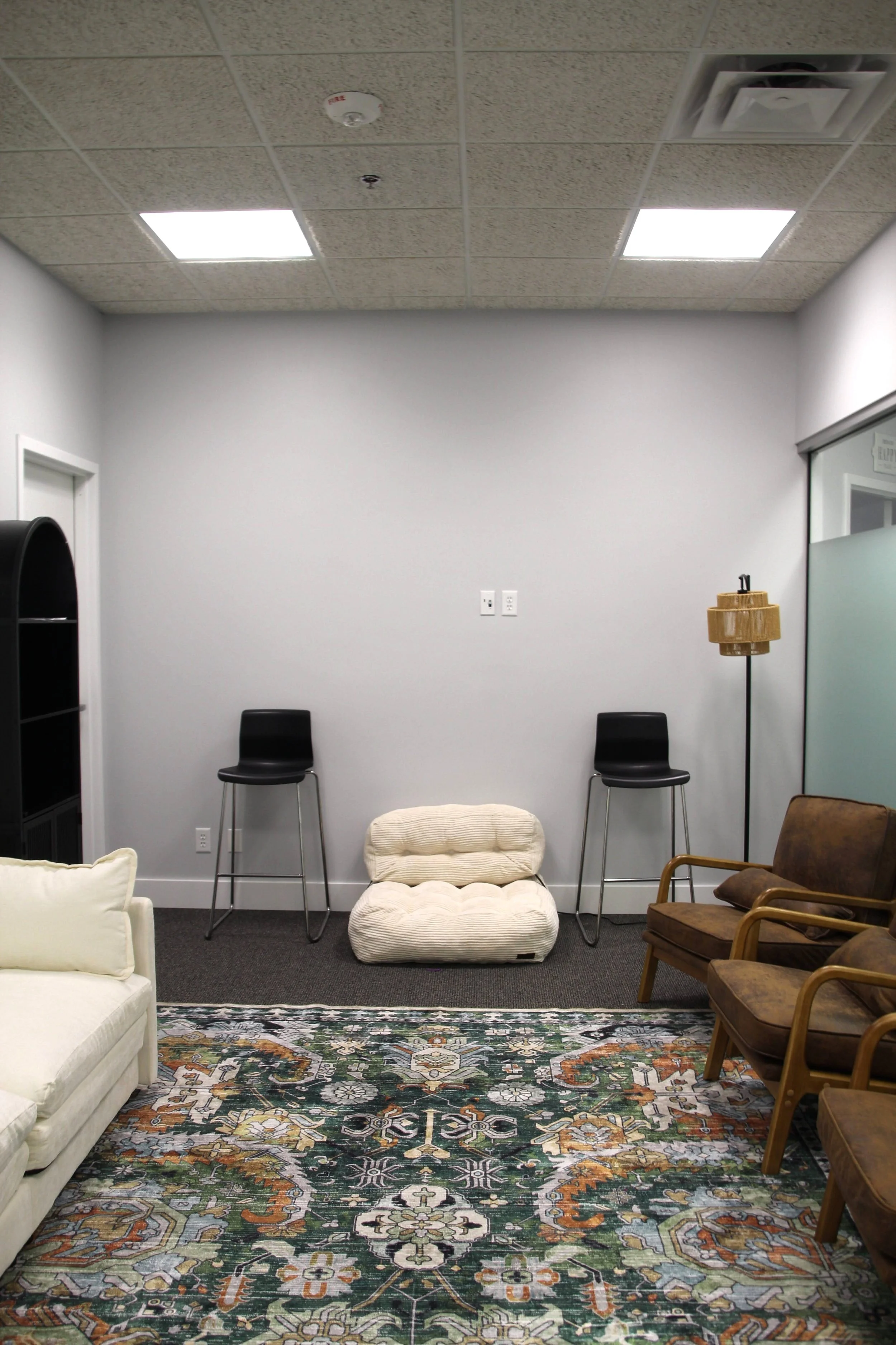 Group therapy room with black and brown chairs, a white sofa, patterned green and orange rug, and a white wall with two black chairs and a small vintage floor lamp.
