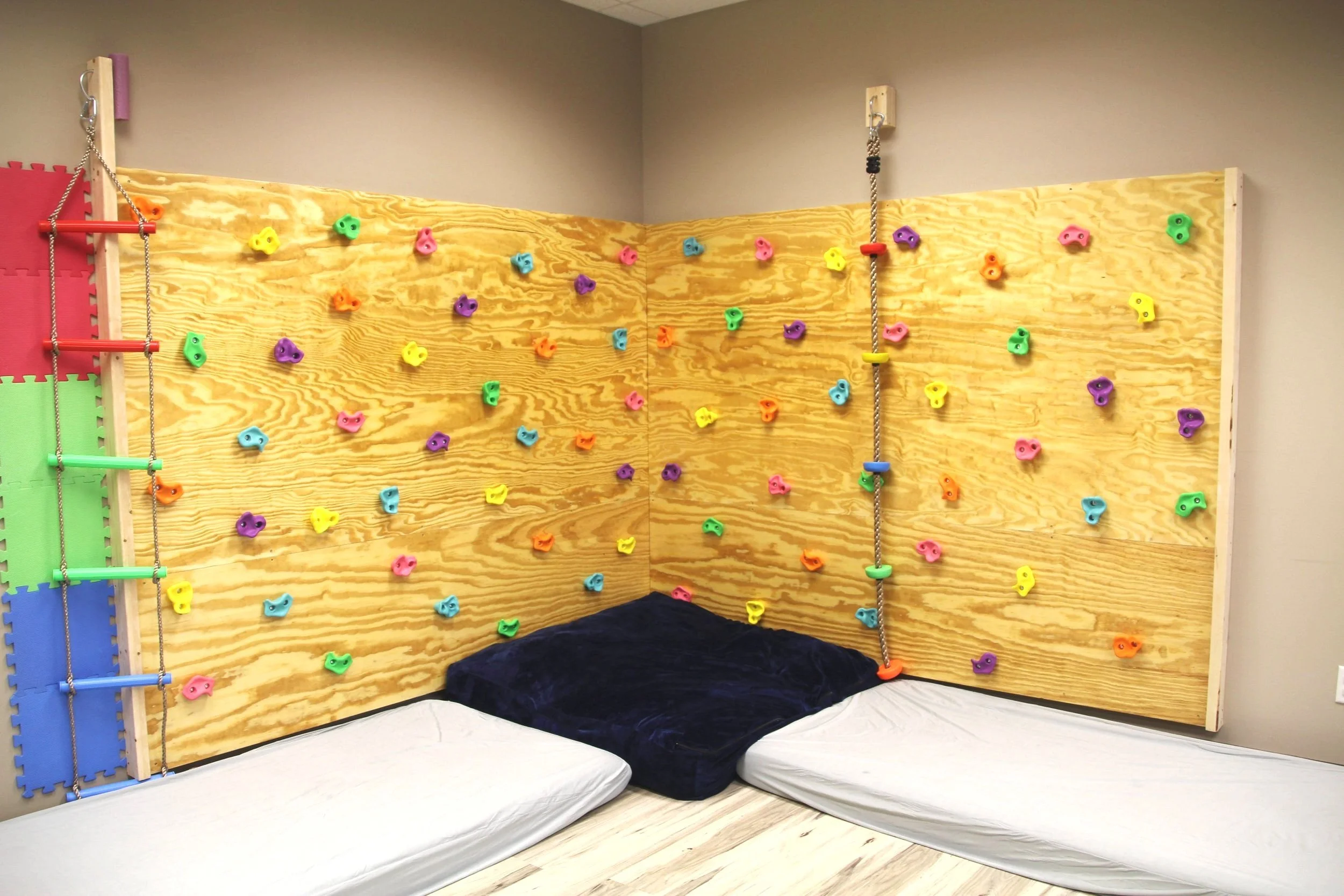 Indoor climbing wall with colorful holds on wooden panels, padded mats on the floor, and a climbing rope.