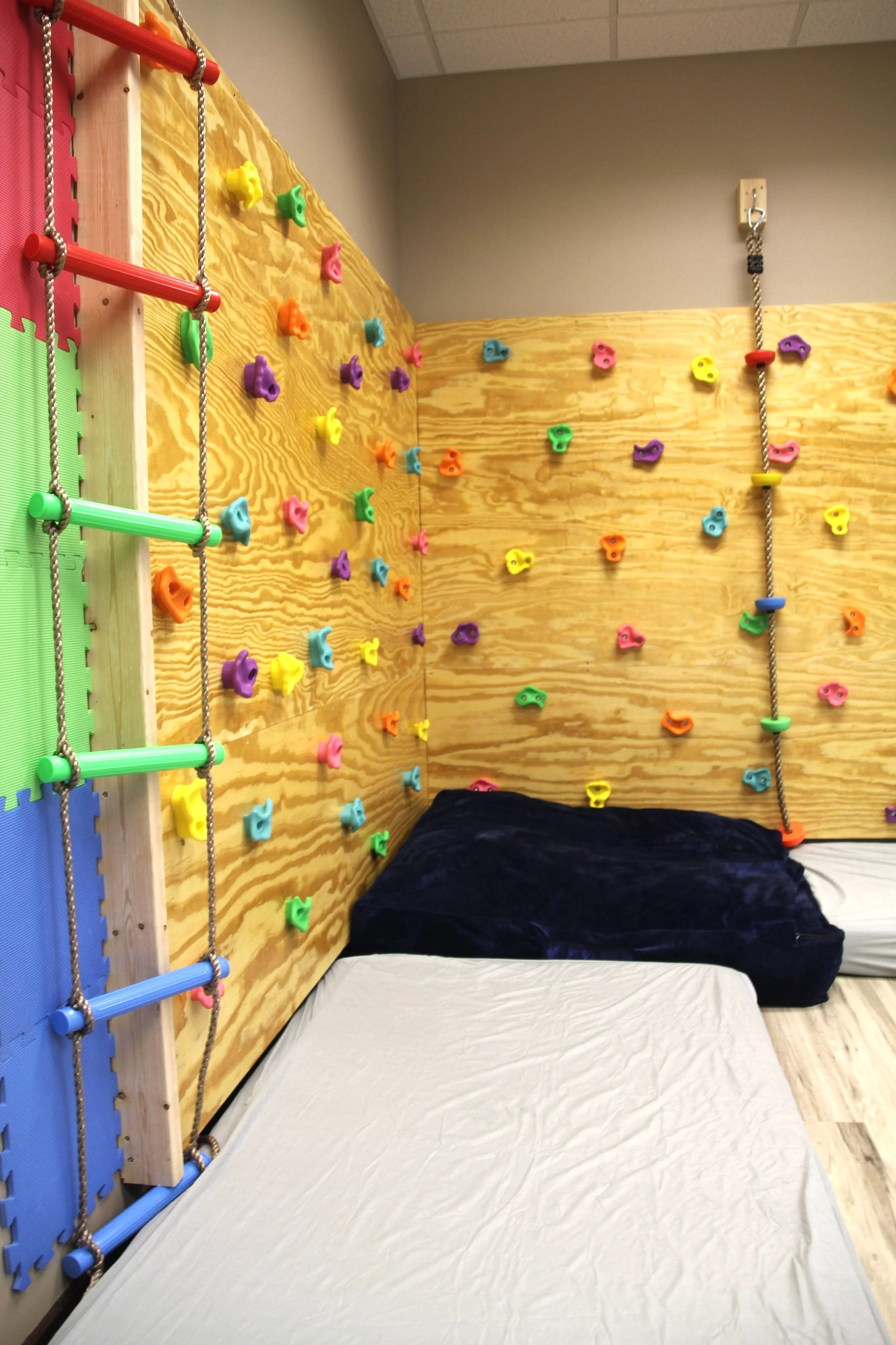 Indoor play area with a climbing wall decorated with colorful holds, a climbing rope, foam mats, and a cushioned black mat for safety.